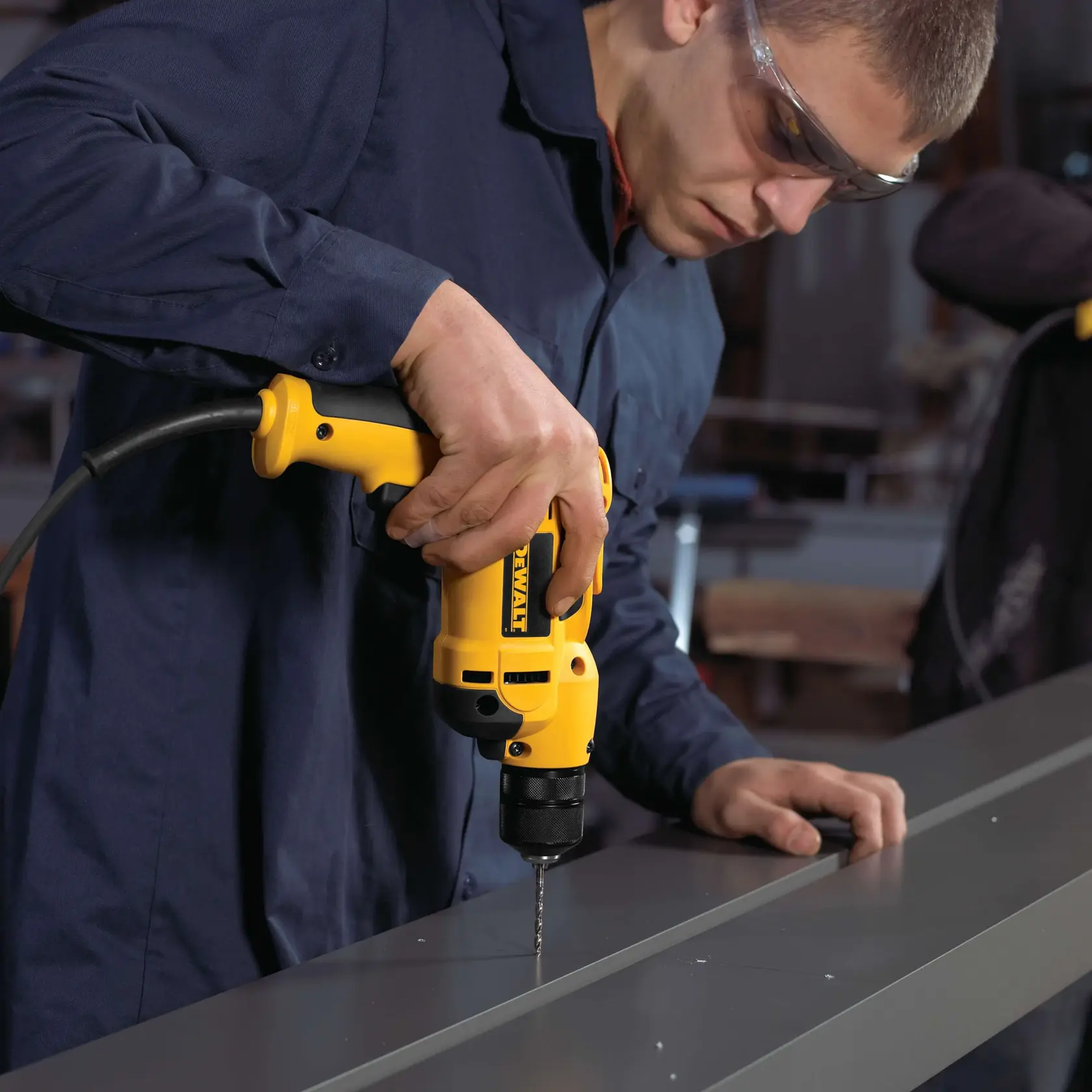 A person using a yellow DEWALT DWD112 electric drill to create holes in a metal surface. The person is wearing a dark blue work shirt and their face is blurred for privacy.