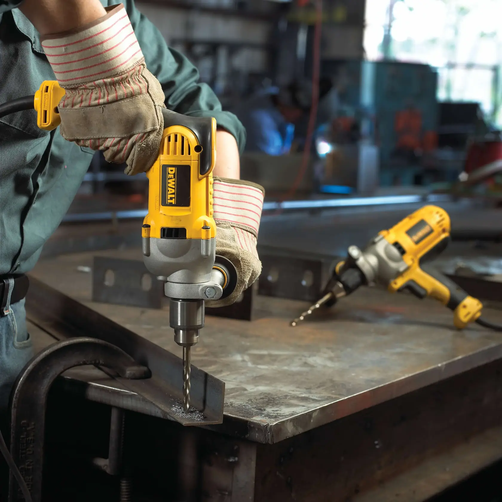 A person wearing work gloves is using a yellow DEWALT electric drill to bore a large hole through a wooden stud inside a construction frame. The drill features a long auger bit and there is a blue electrical outlet box mounted nearby.
