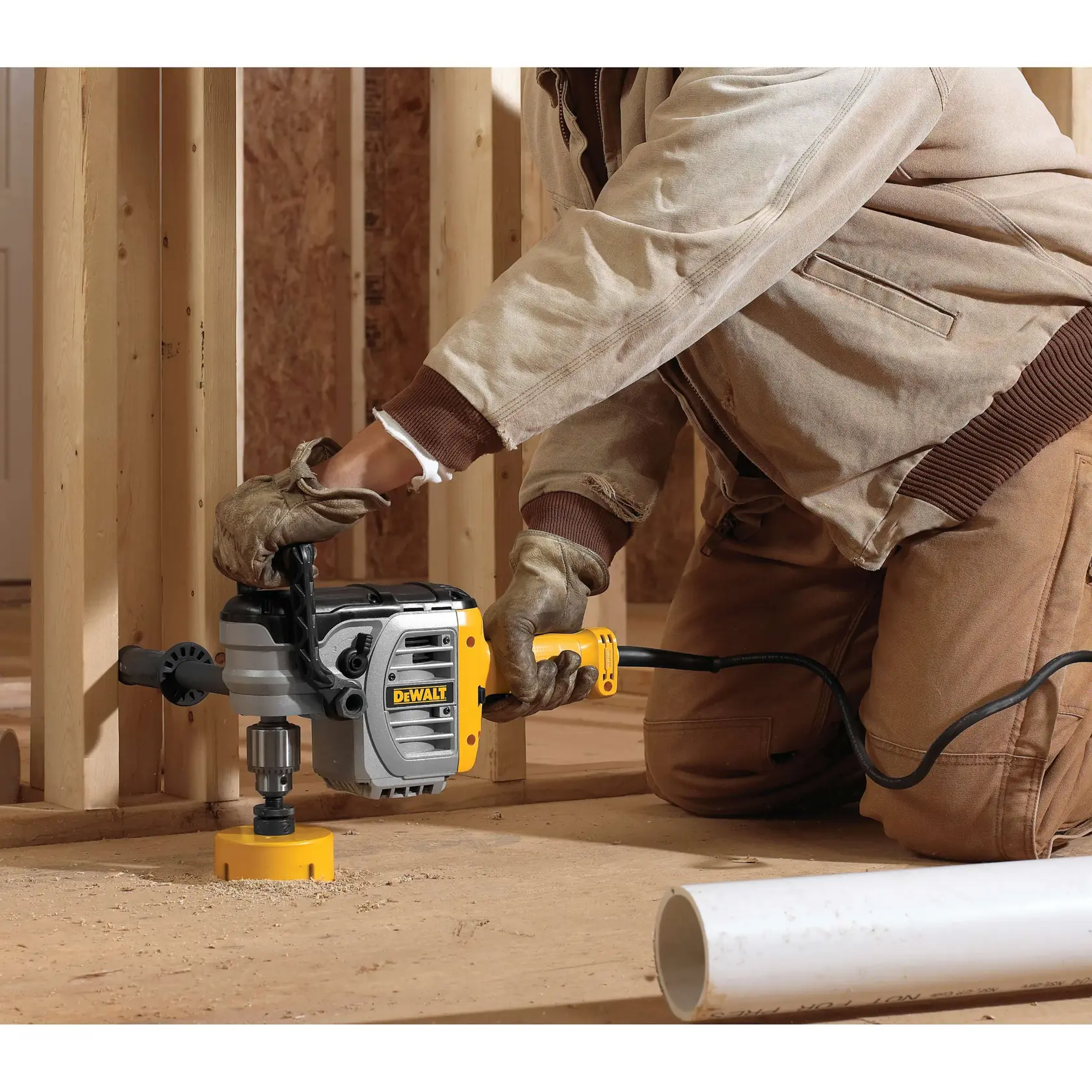 A person using a DEWALT DWD450 power drill with a hole saw attachment to drill through a wooden floor at a construction site. The person is wearing work gloves and a tan jacket, kneeling next to wooden framing and a white PVC pipe.