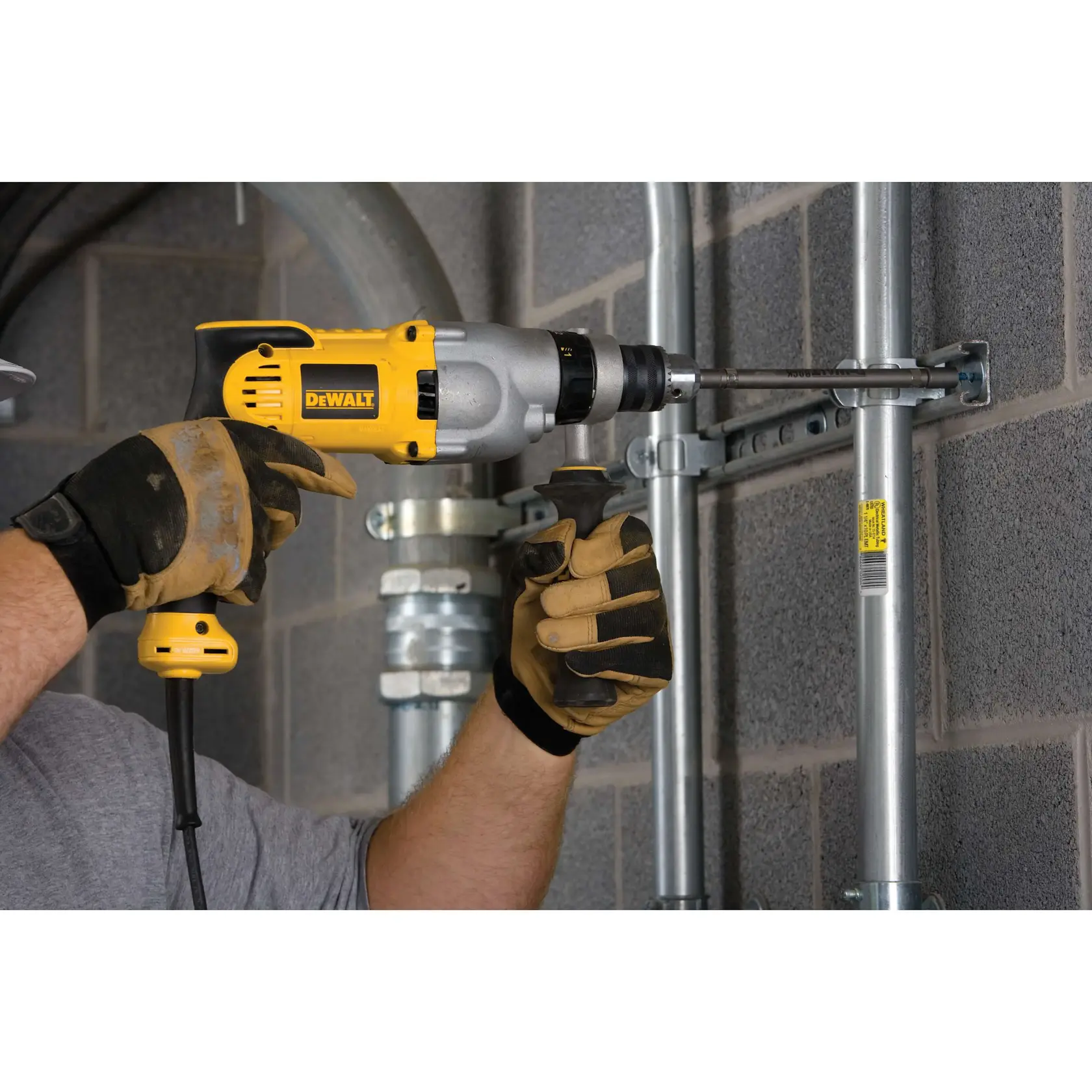 A person wearing work gloves uses a yellow and gray DEWALT DWD520 corded drill to drill into a metal fixture mounted on a concrete wall.