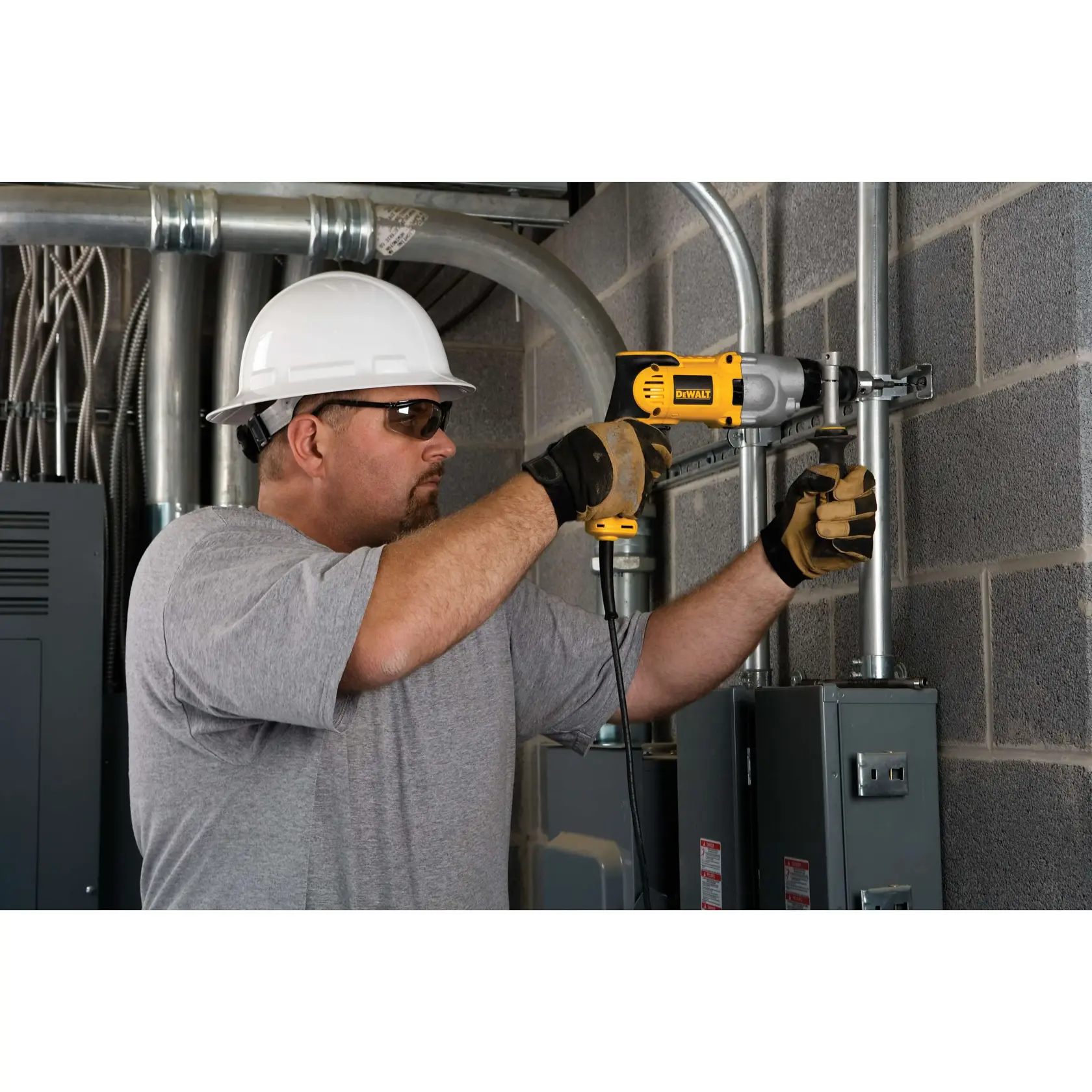 A person wearing a white hard hat and gloves is using a DEWALT DWD520 power drill to work on metal pipes attached to a brick wall in an industrial setting.