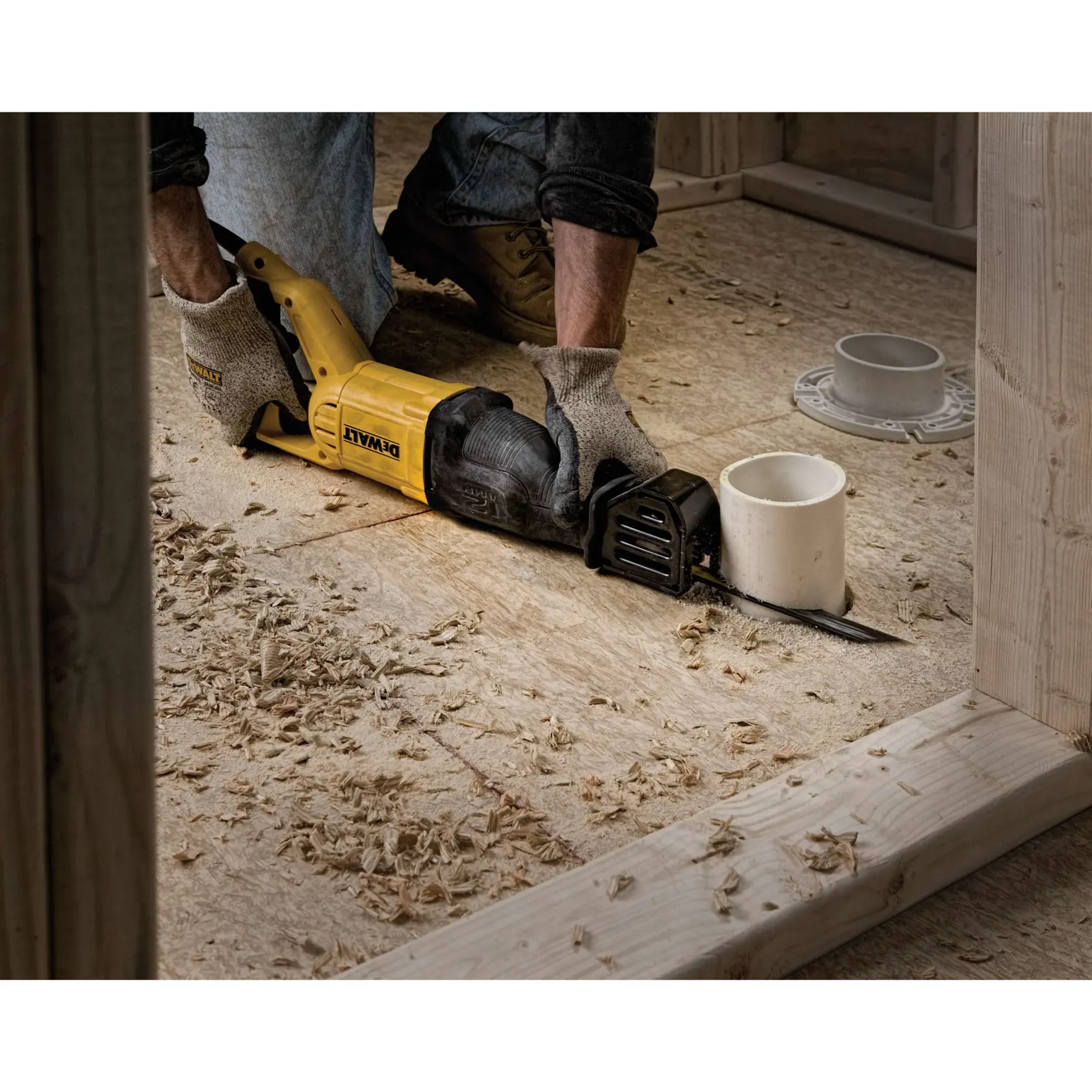A person wearing work gloves uses a yellow DEWALT reciprocating saw to cut through a piece of wood flooring, surrounded by wood shavings and an unfinished construction site.