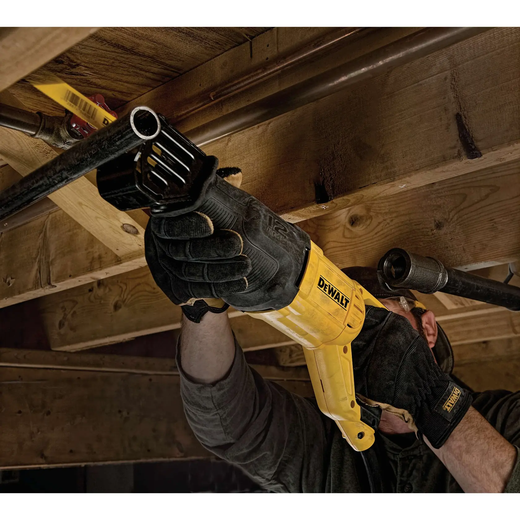 A person wearing gloves uses a yellow DEWALT reciprocating saw (model DWE305) to cut through a metal pipe beneath a wooden ceiling.