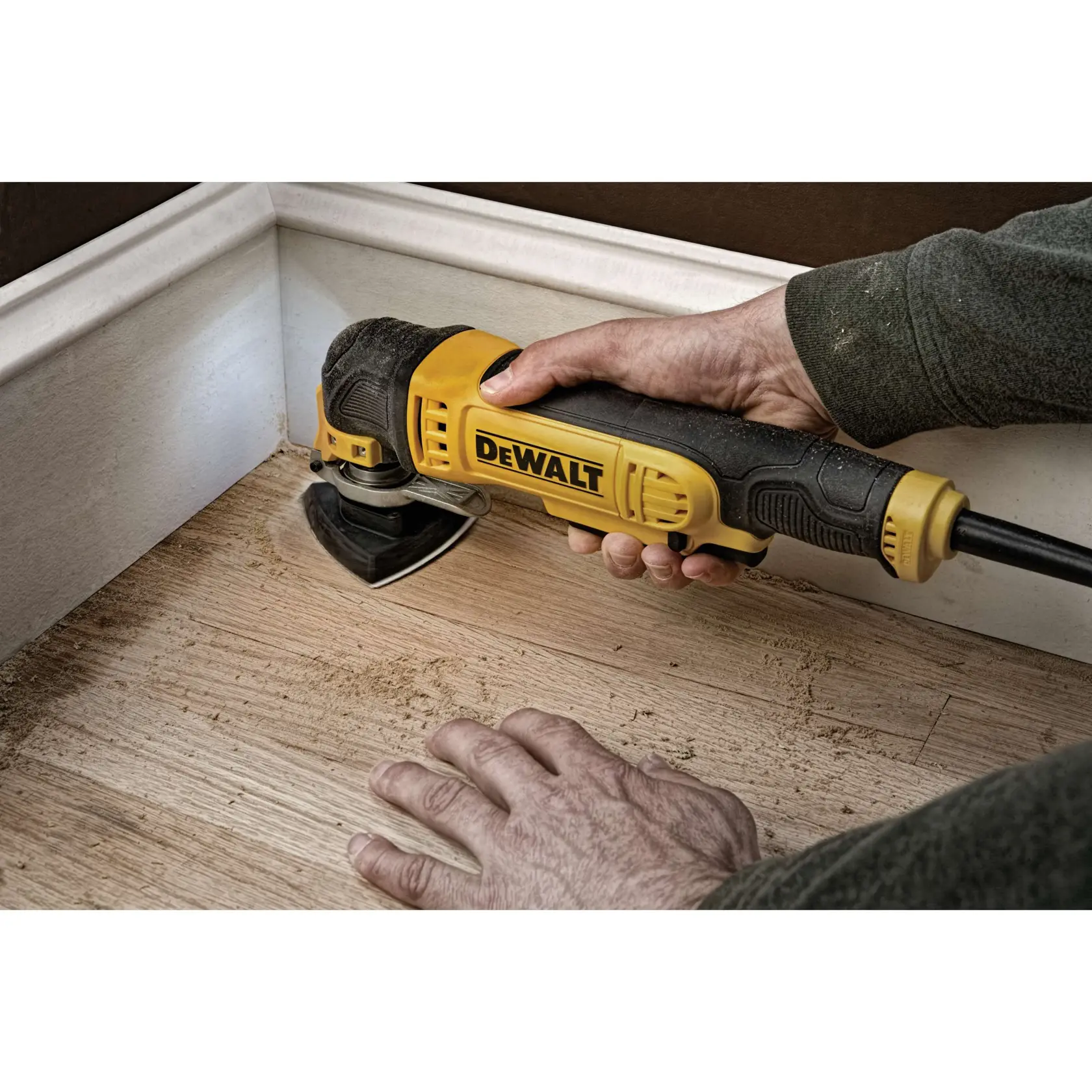 A person is using a DEWALT oscillating multi-tool to sand or cut wood flooring near a baseboard.