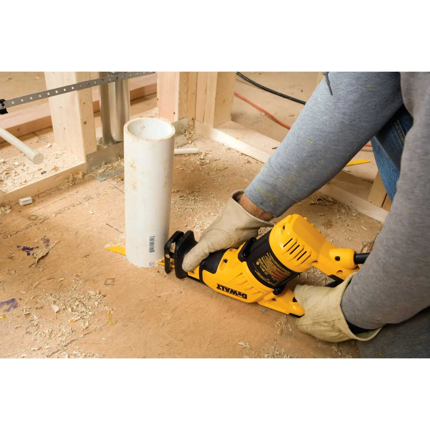 A person wearing gloves uses a yellow DEWALT reciprocating saw to cut a white PVC pipe on a construction site with wood framing and sawdust on the floor.