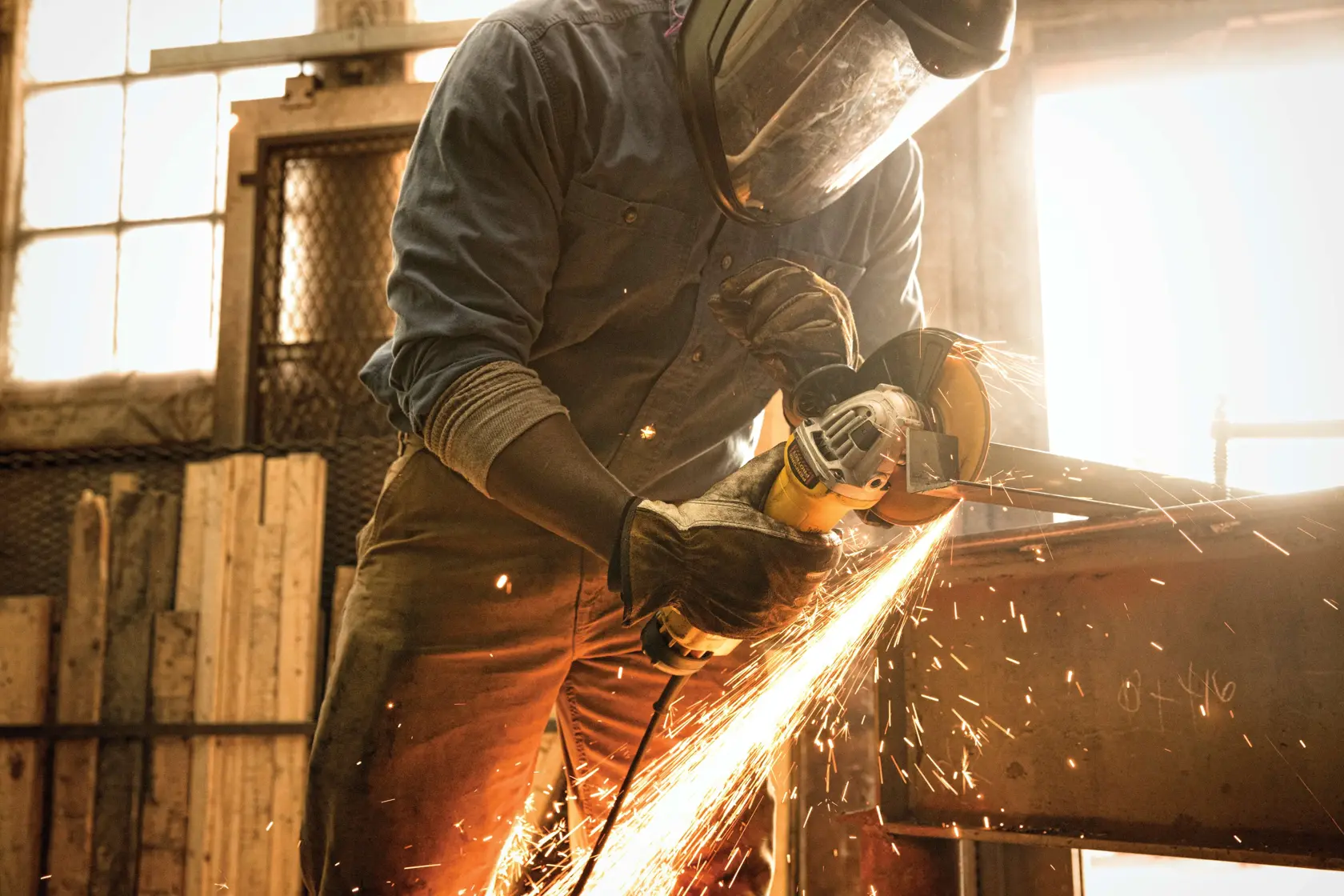 A person wearing protective gear uses a DEWALT angle grinder, producing sparks as they cut or grind a piece of metal in an industrial workshop setting.