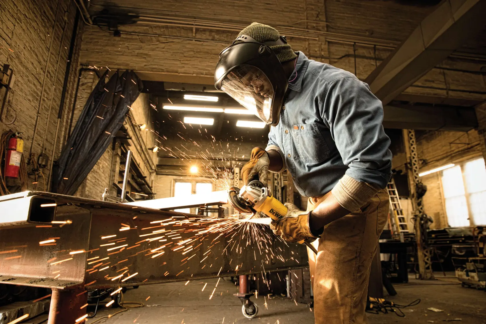 A worker wearing protective gear, including a face shield and gloves, uses a DEWALT angle grinder to cut metal in an industrial workshop, with sparks flying from the grinding process.