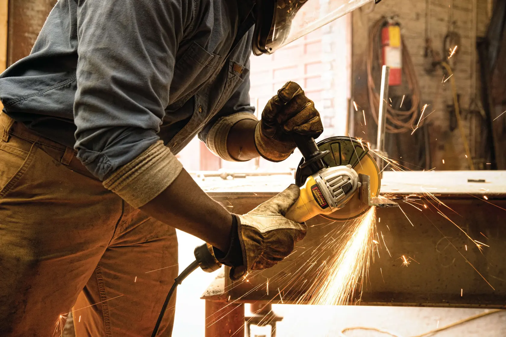 A person wearing protective gloves and a face shield is using a DEWALT angle grinder, generating sparks while working on a metal surface in an industrial workshop.
