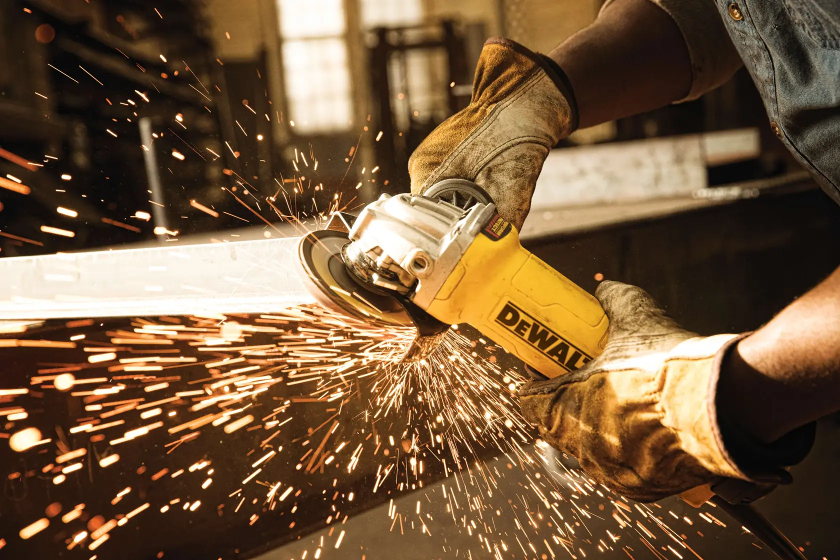 A person wearing protective gloves is using a DEWALT angle grinder to cut metal, with sparks flying in a workshop environment.