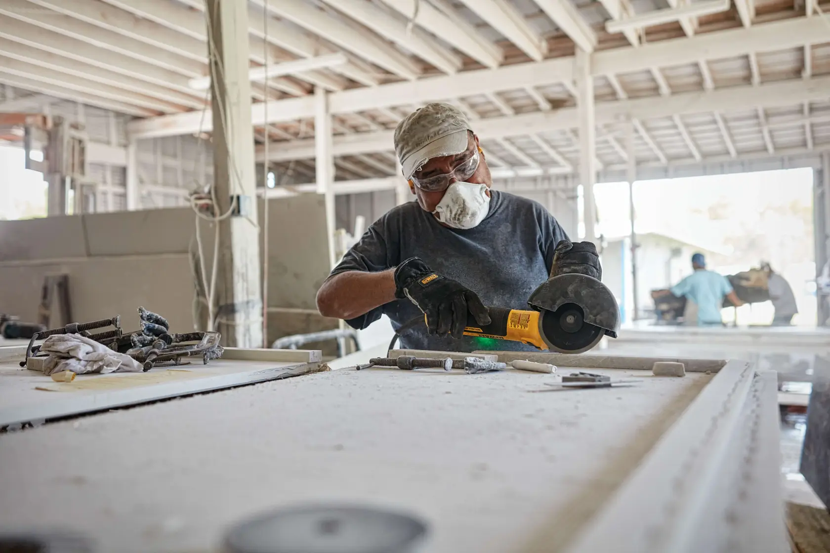 Person wearing a cap and gloves using a DEWALT DWE43240VS angle grinder on a dusty worktable in an industrial workshop.