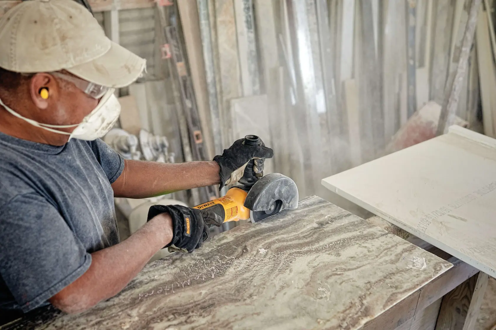A person wearing a cap, ear protection, gloves, and a mask is using a yellow DEWALT angle grinder to cut or polish a stone slab in a workshop filled with various construction materials.