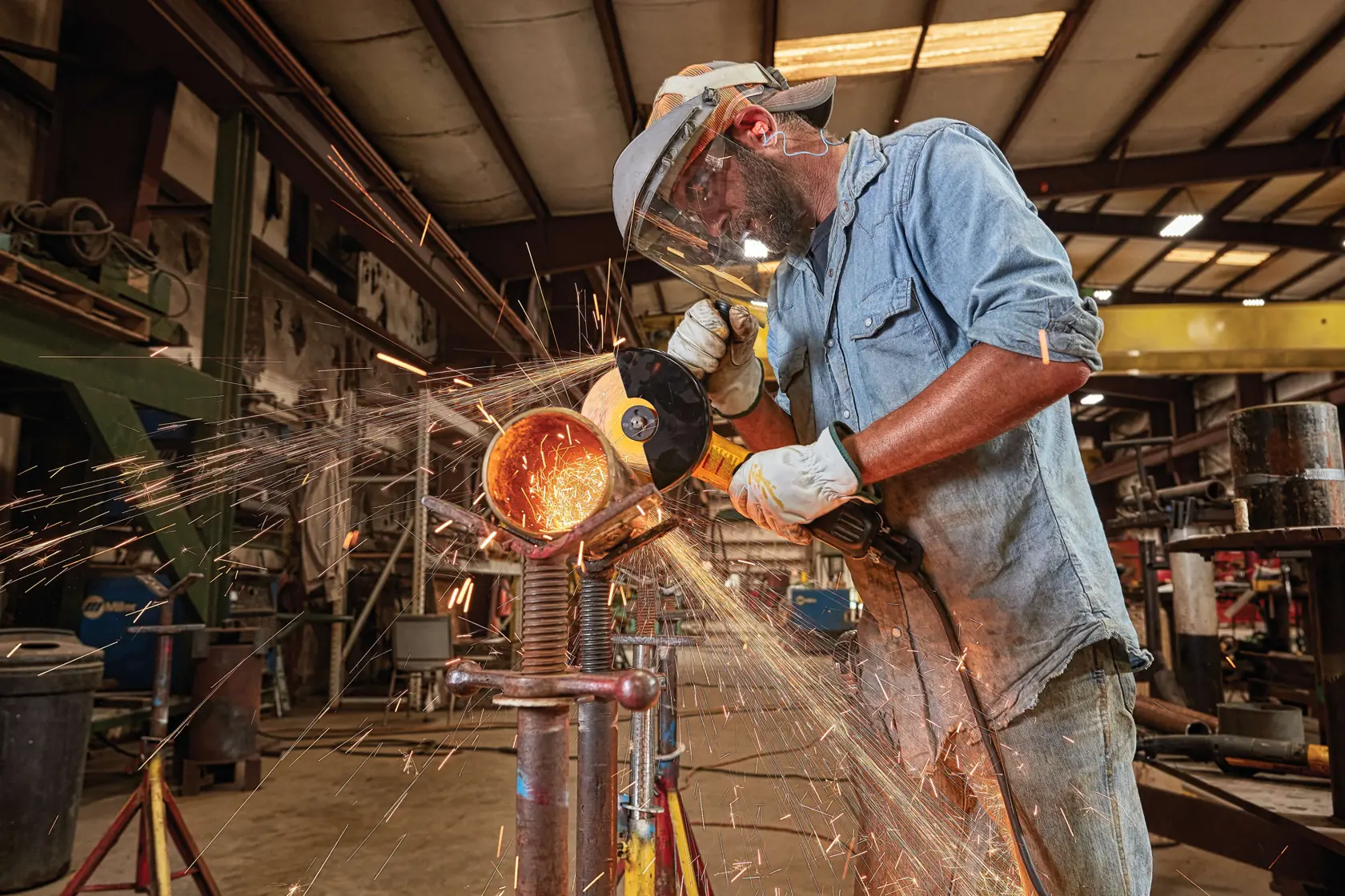 A person wearing protective gear uses a DEWALT angle grinder to cut a metal pipe, producing sparks in an industrial workshop setting.