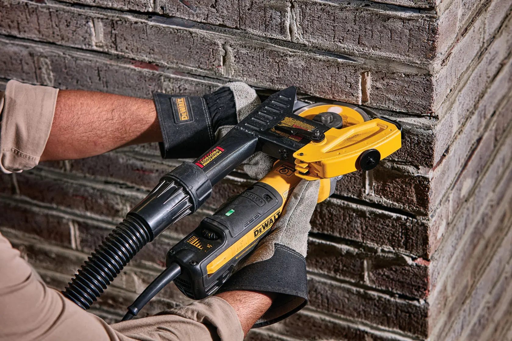A person wearing gloves uses a DEWALT electric power tool, equipped with a dust extraction attachment, to cut or grind mortar joints on a brick wall corner.