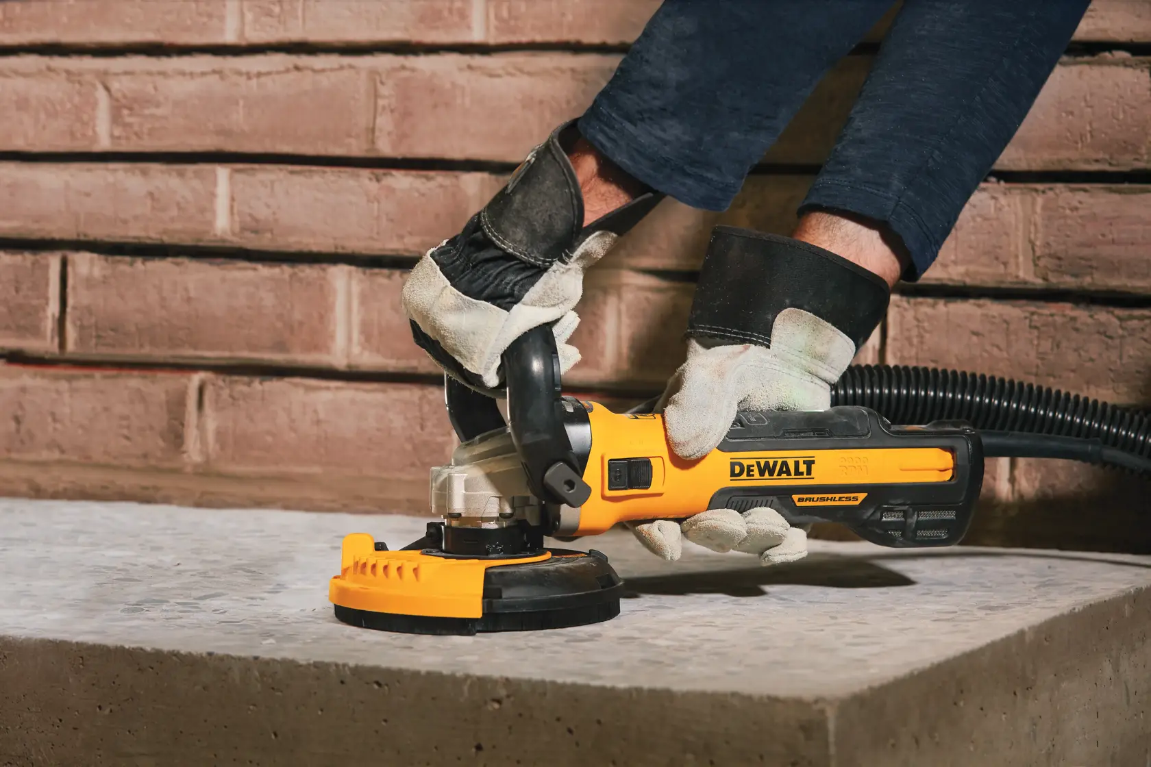A person wearing work gloves uses a DEWALT electric grinder on a concrete surface. The grinder is yellow and black with a dust extraction attachment. The person is working near a brick wall.