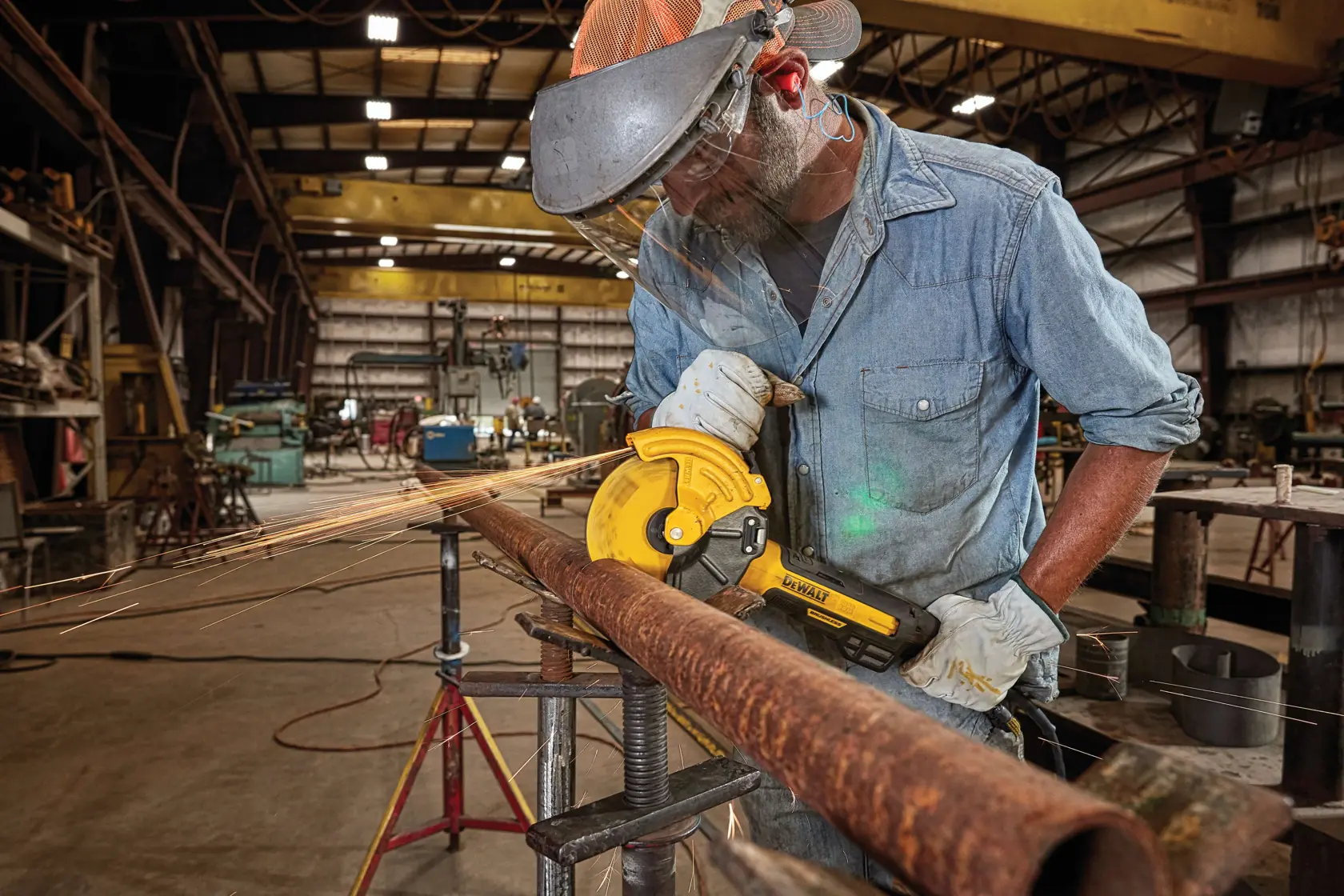 A person using a DEWALT angle grinder to cut a rusty metal pipe in an industrial workshop. The person is wearing protective gear including gloves, a face shield, and ear protection.