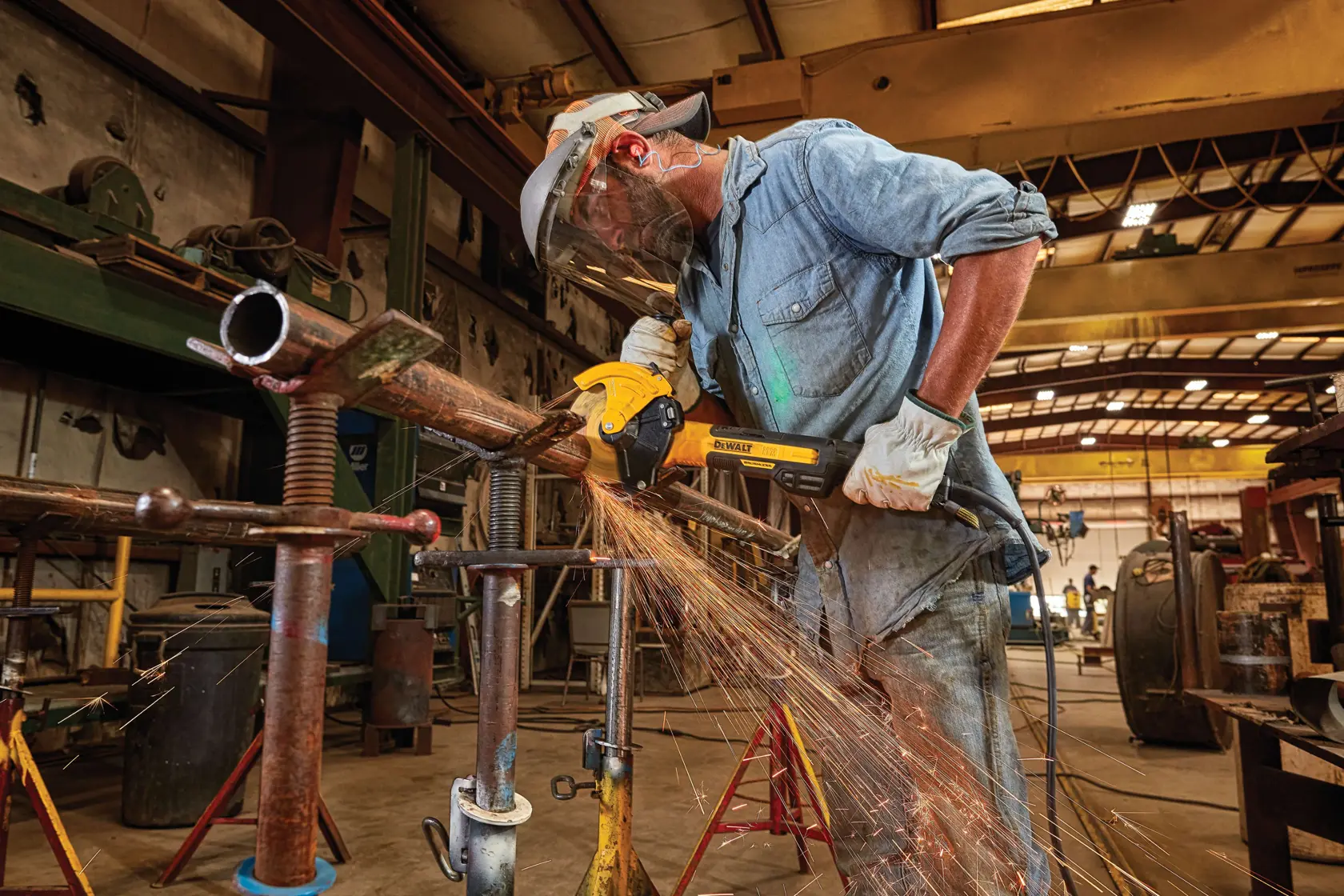 A worker in protective gear uses a DEWALT angle grinder to cut a metal pipe, producing sparks, in an industrial workshop setting.
