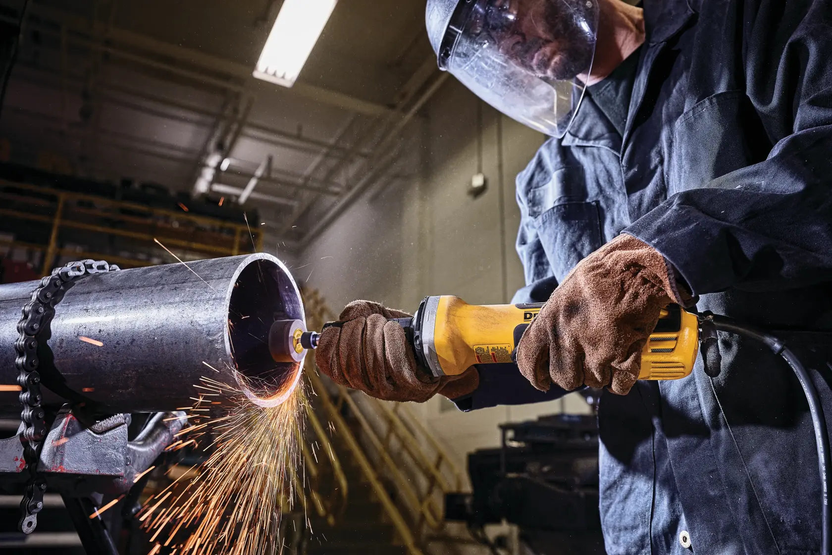 A person wearing gloves and a protective face shield is using a yellow DEWALT angle grinder to cut or grind the edge of a large metal pipe, producing sparks in an industrial workshop setting.