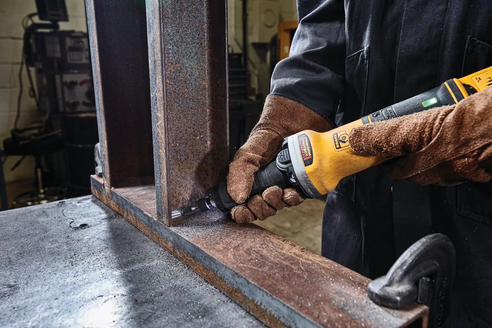 A person wearing brown protective gloves is using a yellow DEWALT power tool to grind or polish the edge of a large, rusty metal beam in an industrial workshop setting.