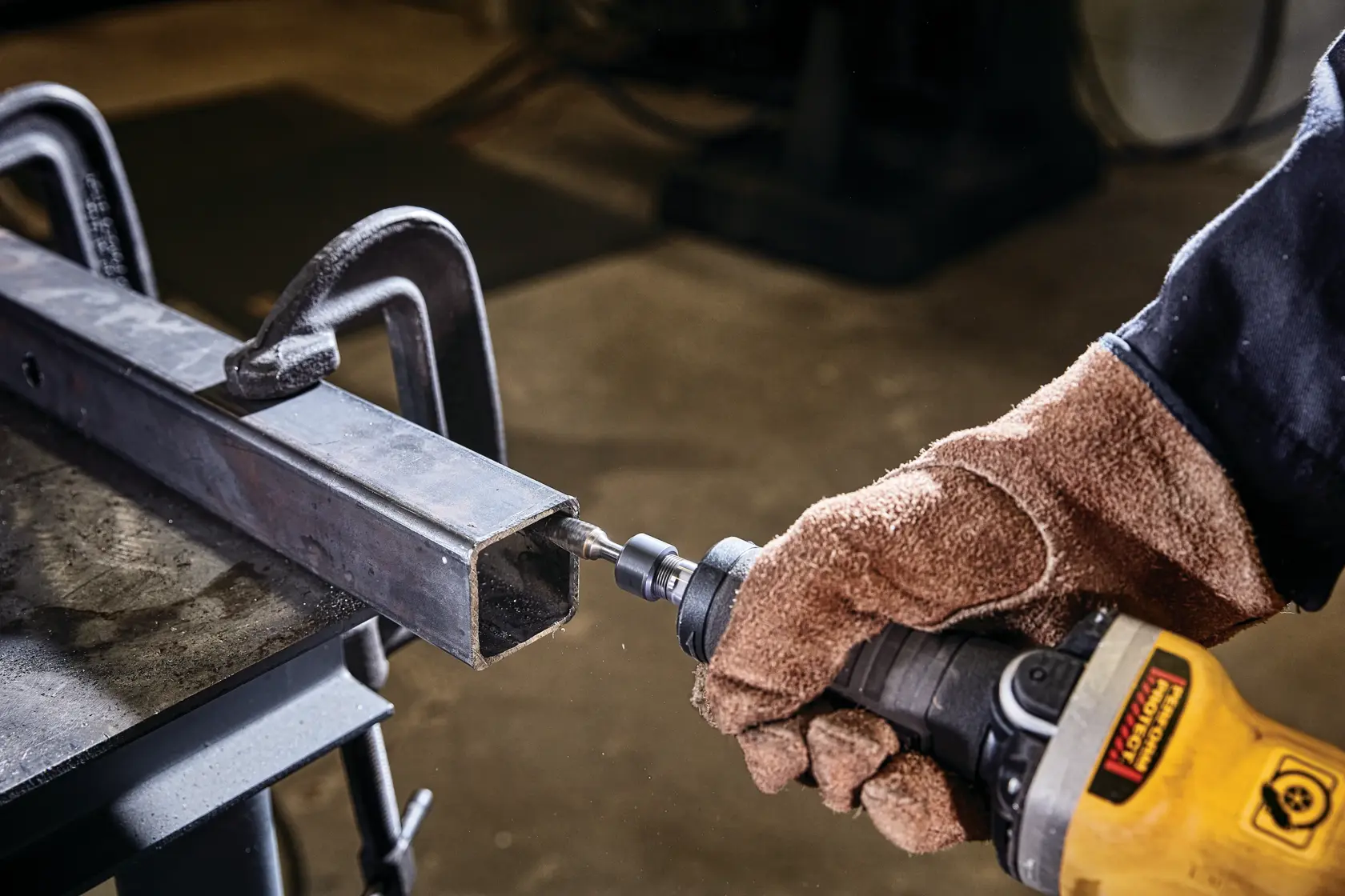 A person wearing protective gloves uses a DEWALT power tool to grind or polish the inside edge of a metal square tube, which is clamped securely to a workbench.