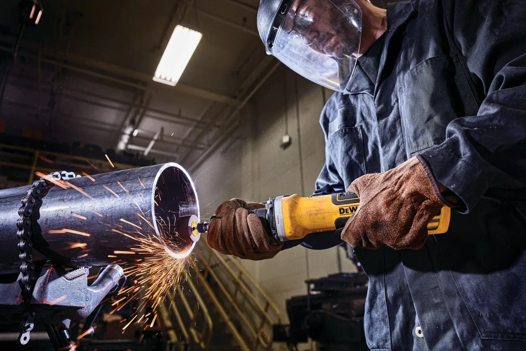 A person wearing protective gloves and a face shield is using a yellow DEWALT power tool to grind a metal pipe, with sparks flying in an industrial workshop setting.