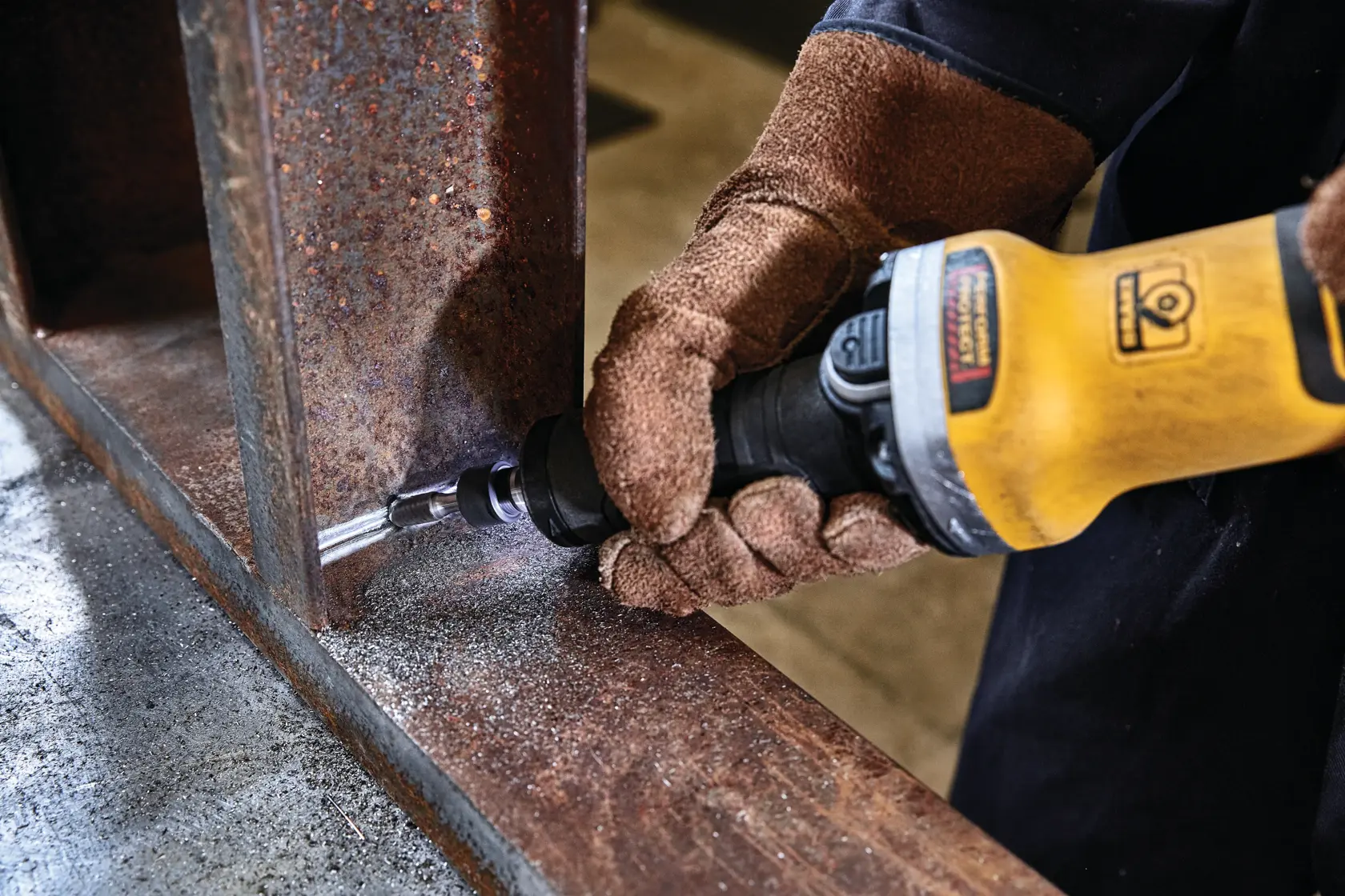 A person wearing brown work gloves uses a yellow DEWALT power tool to grind or polish the inside corner of a rusty metal beam.