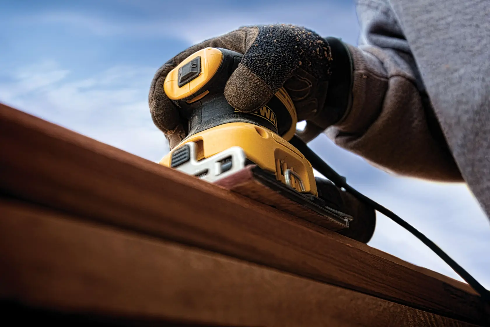 A close-up of a gloved hand using a DEWALT electric sander on a wooden surface outdoors, with a blue sky in the background.