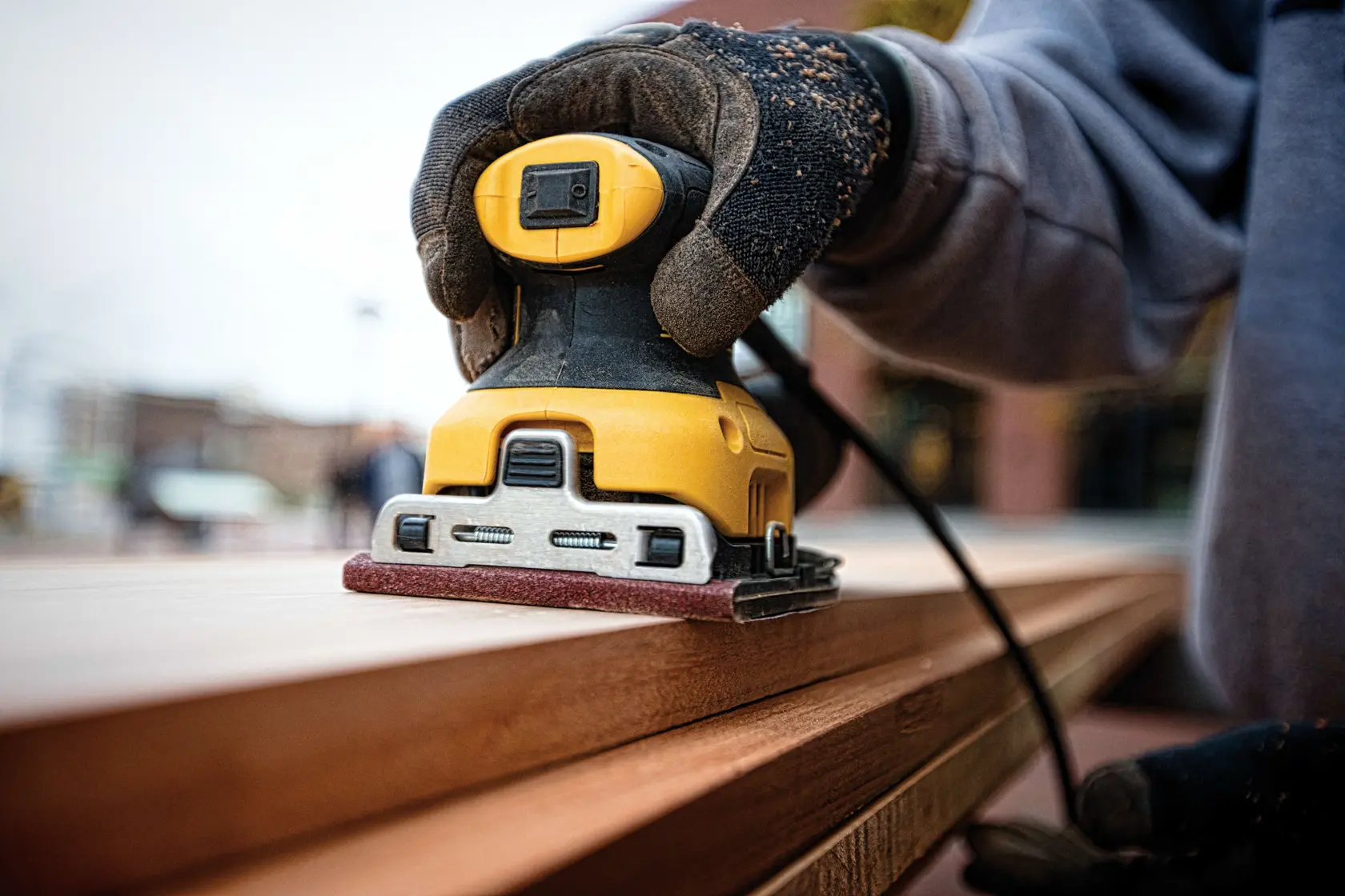 A close-up of a person wearing gloves using a yellow DEWALT electric sander on a wooden surface outdoors.