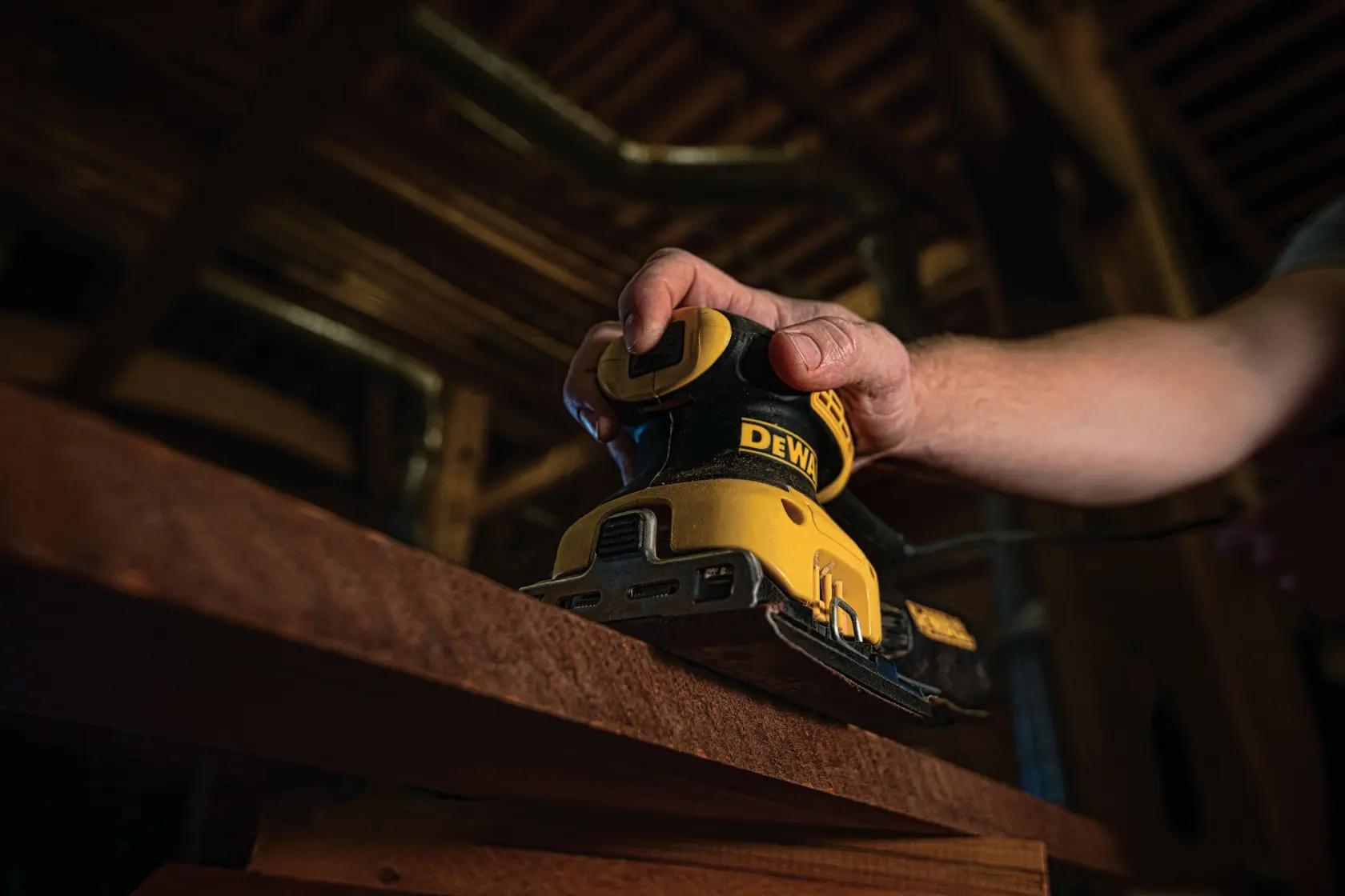 A close-up view of a person's hand using a DEWALT DWE6411K electric sander on a wooden surface in a workshop setting.