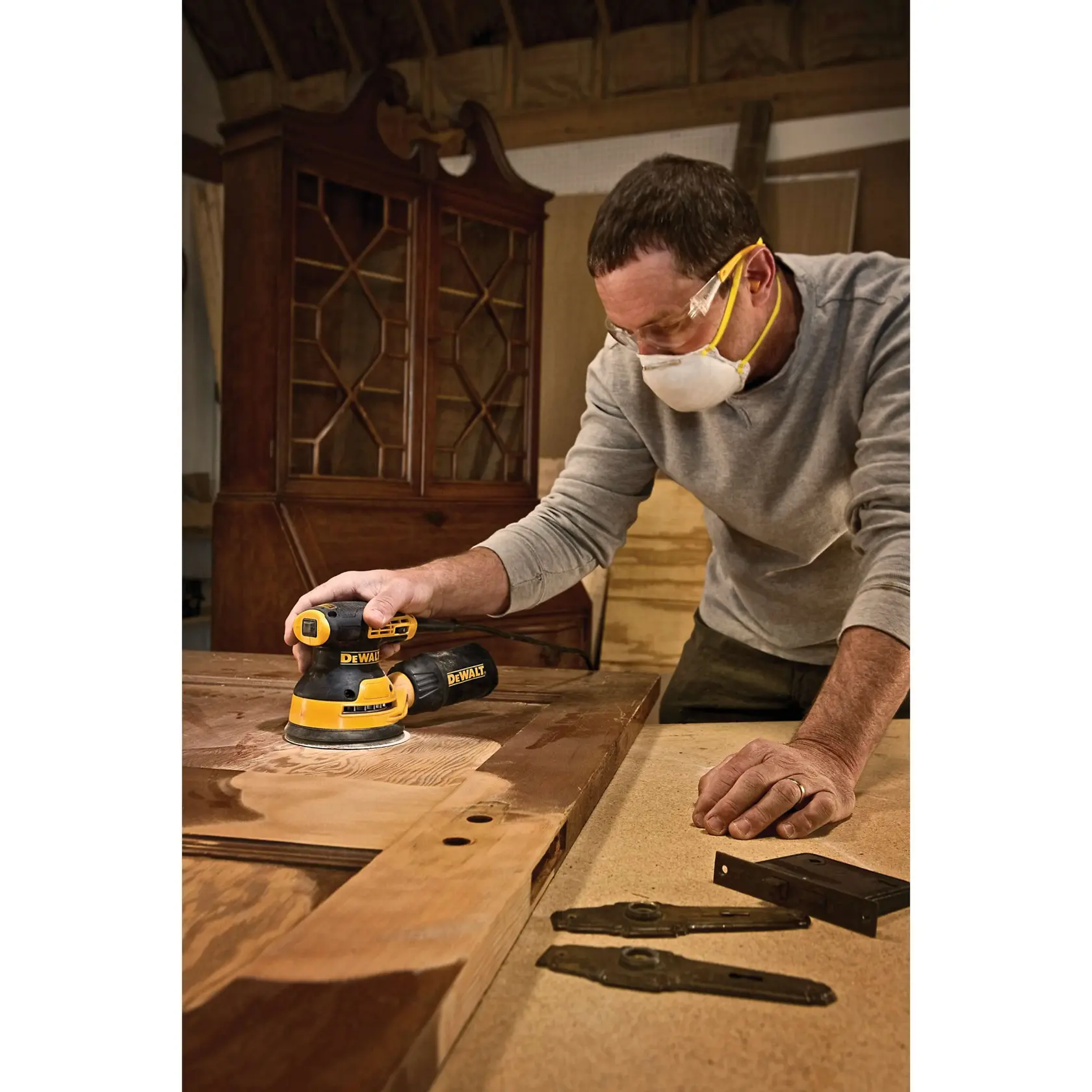 A person using a DEWALT DWE6423K random orbit sander to smooth a wooden door on a workbench. The sander is yellow and black, and the person is working in a woodworking shop. Their face is blurred for privacy.