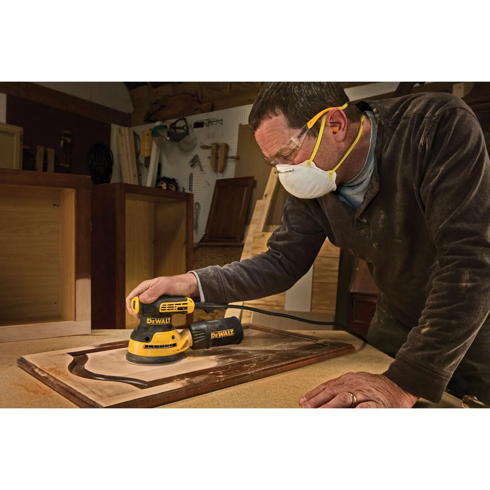 Person using a DEWALT electric sander (model DWE6423K) to smooth a wooden cabinet door in a workshop. The person is wearing safety goggles and a dust mask for protection.