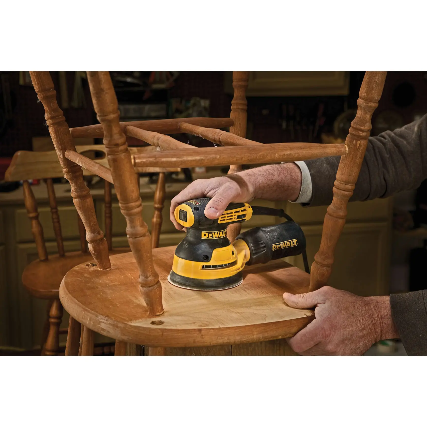 A person is using a DEWALT DWE6423K random orbital sander to sand a wooden chair in a workshop.
