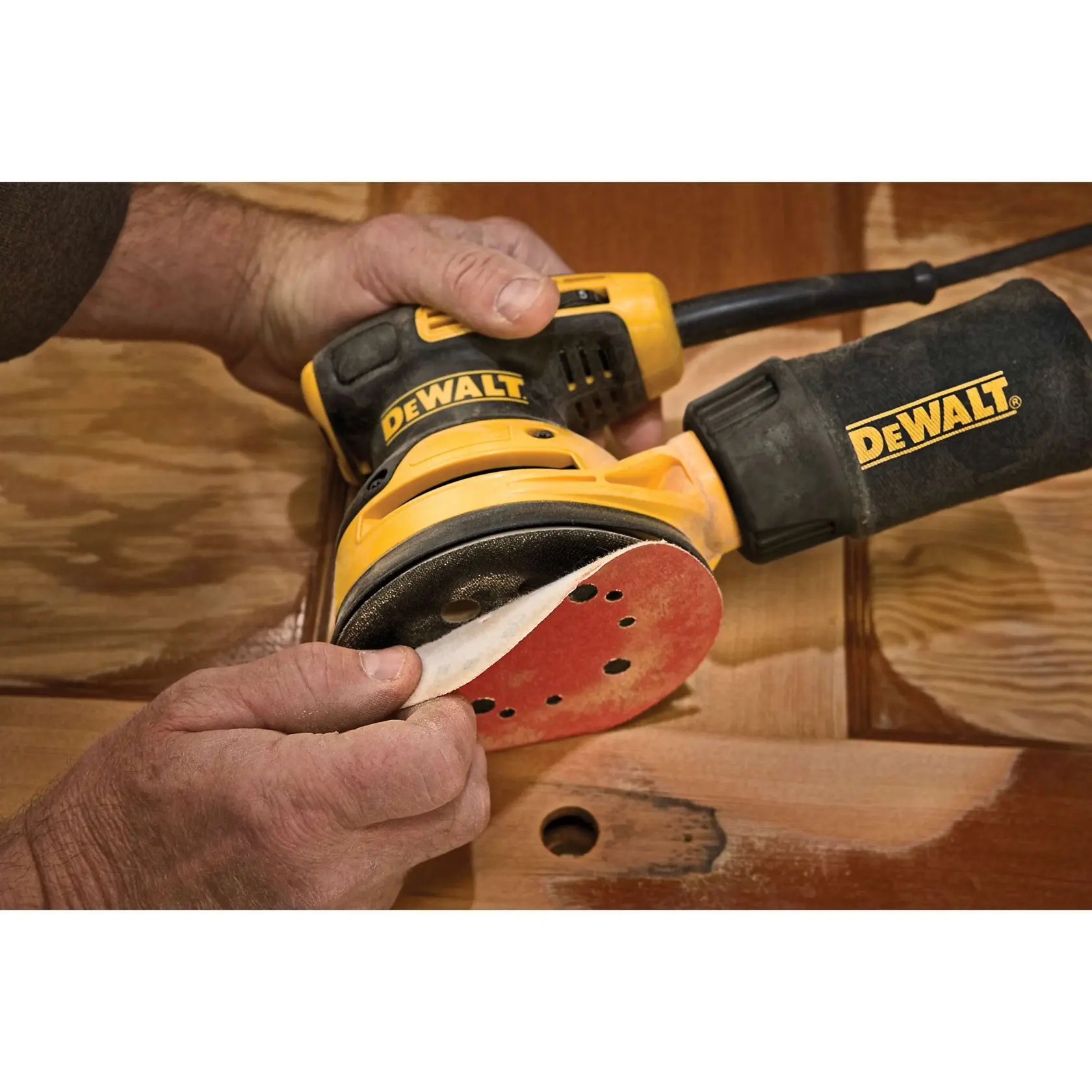 Close-up of hands attaching a sanding disc to a DEWALT random orbit sander, with the tool resting on a wooden surface.