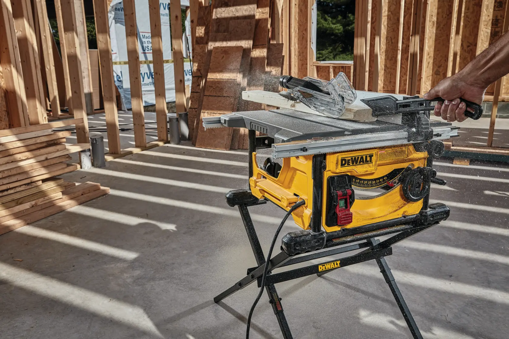 A DEWALT portable table saw is cutting a wooden plank at a construction site with exposed wooden framing and stacks of lumber nearby.