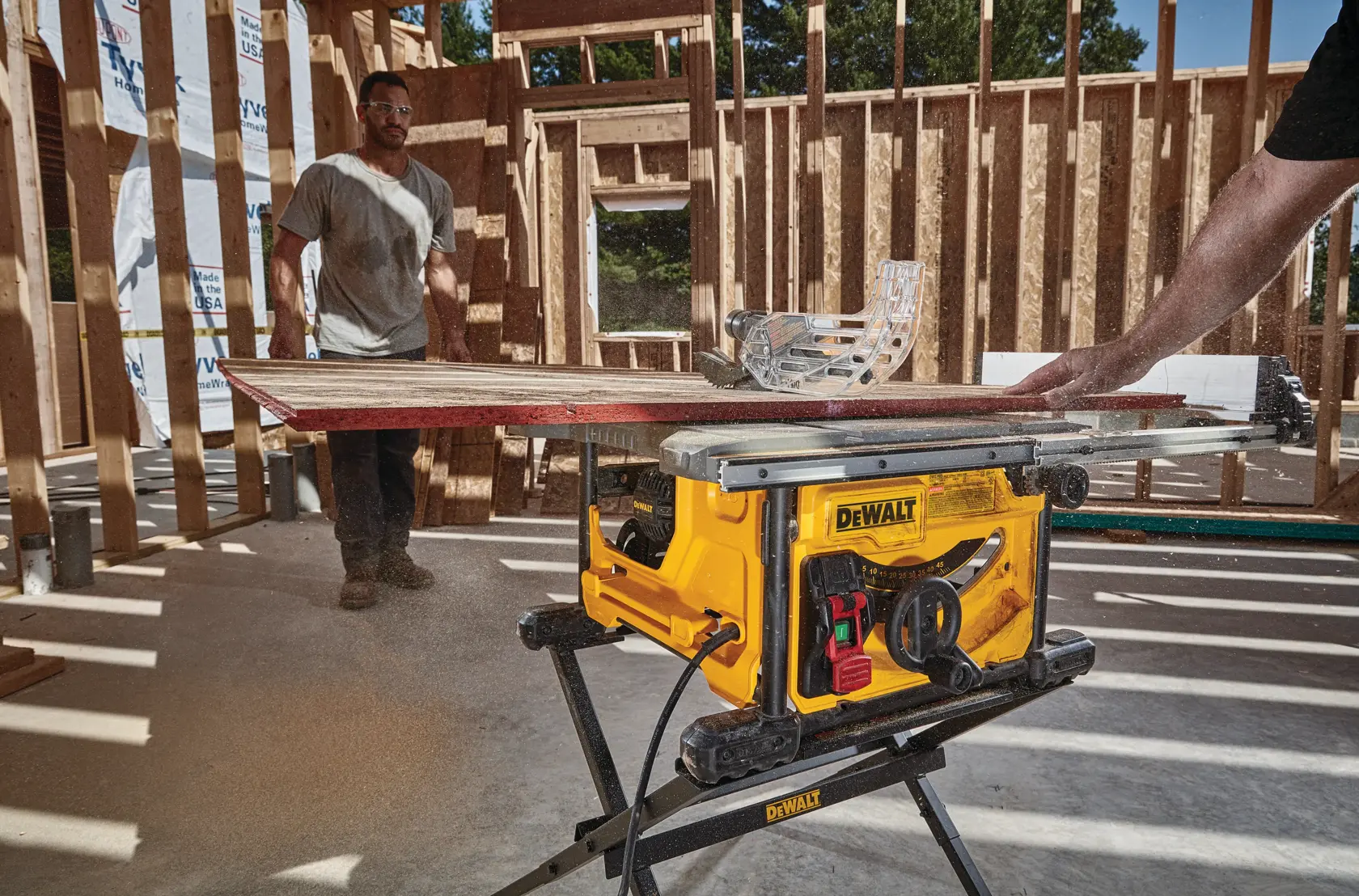A yellow DEWALT table saw is cutting a piece of wood inside a partially constructed wooden building. One person is operating the saw, and another stands in the background. Sawdust is visible around the machine.