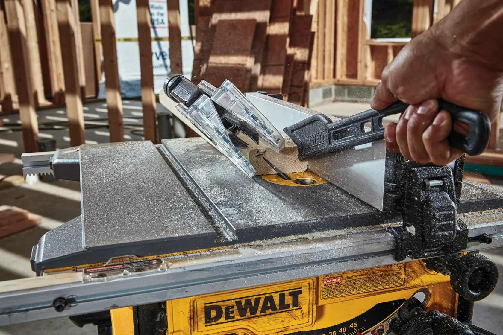 A close-up view of a DEWALT table saw in use, with a person's hand guiding a piece of wood through the cutting blade using a push stick. The saw is being operated at a construction site with wooden framing in the background.