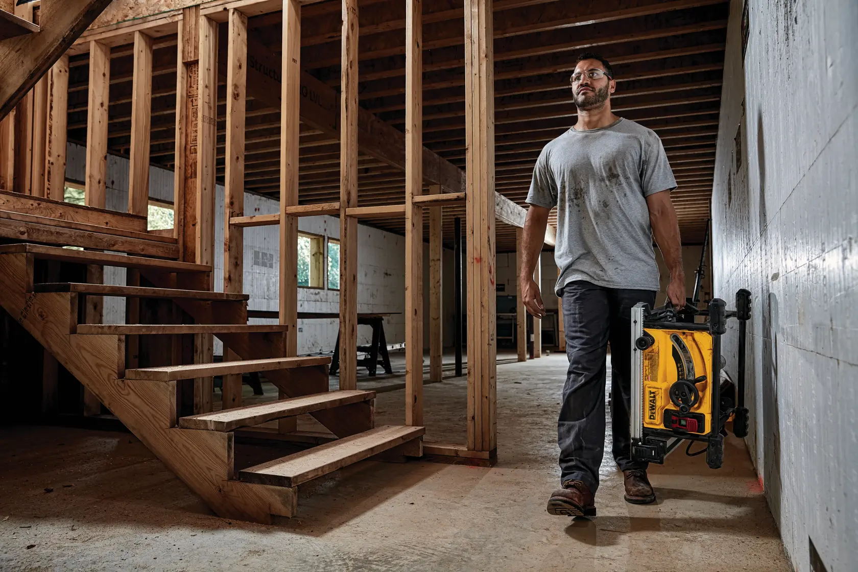 Person carrying a DEWALT portable table saw with stand in a partially constructed building, walking near unfinished wooden stairs and framed walls.