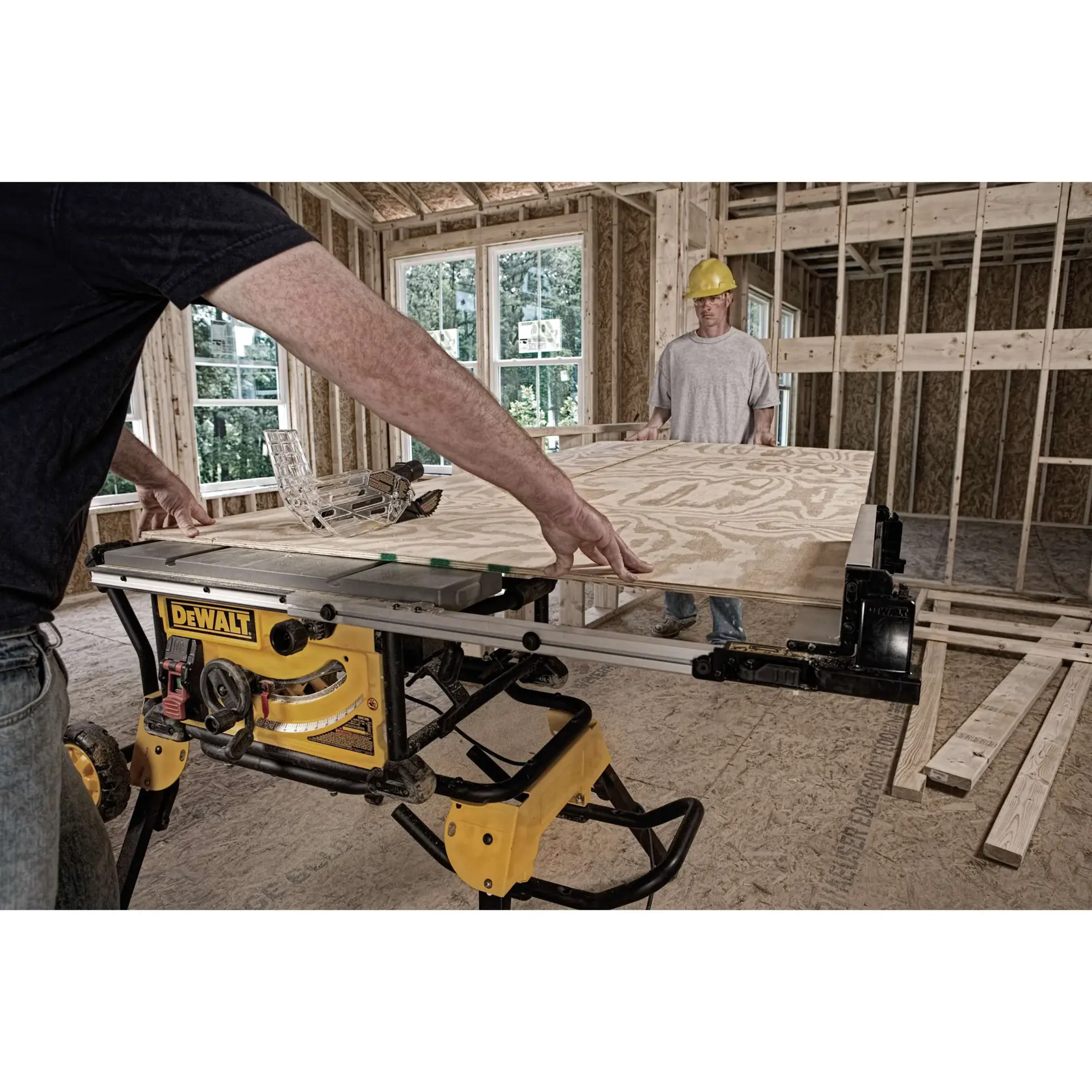 A DEWALT table saw with stand is being used to cut a large sheet of plywood in a construction site. Two people are working together inside a partially built wooden structure; one person is guiding the plywood while the other assists, wearing a safety helmet.