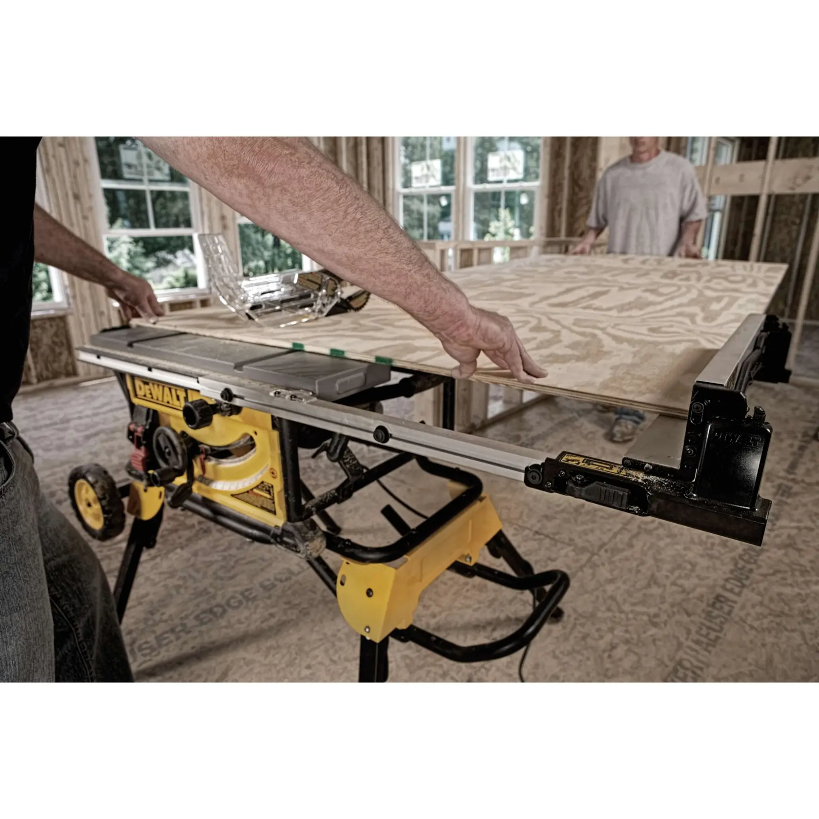 A person is using a DEWALT table saw with a yellow stand to cut a large sheet of plywood in a construction setting. Another person is in the background assisting.