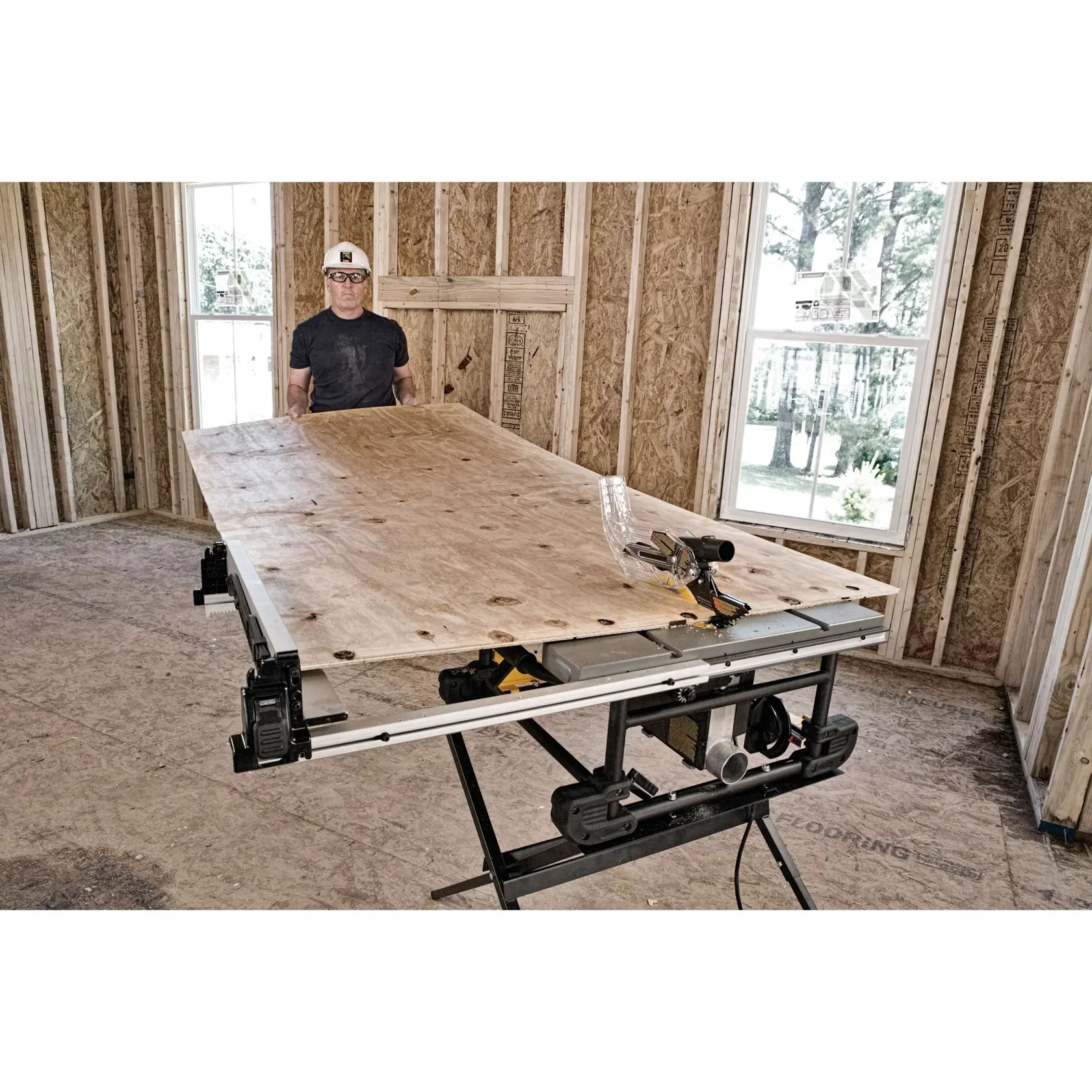 A person is preparing to cut a large sheet of plywood using a DEWALT table saw inside a building under construction, with unfinished walls and visible windows.