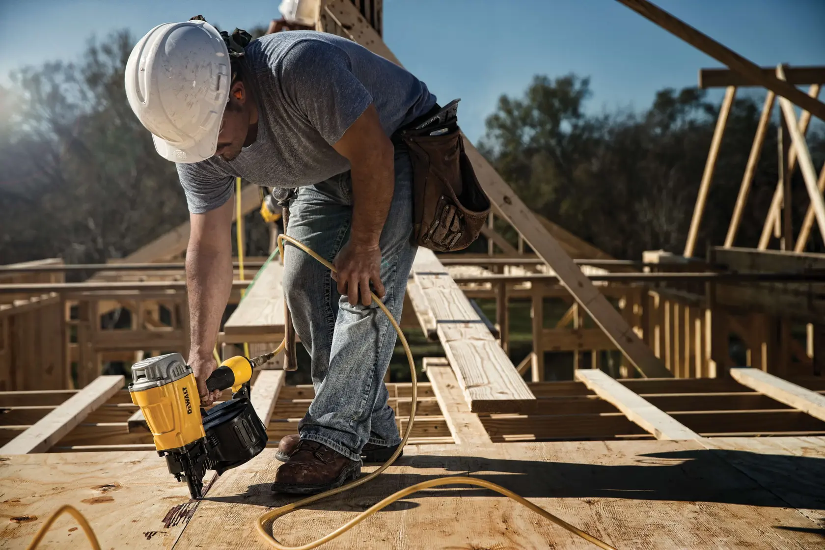 A construction worker wearing a hard hat uses a DEWALT pneumatic nail gun to fasten plywood on a wooden house frame at a building site.