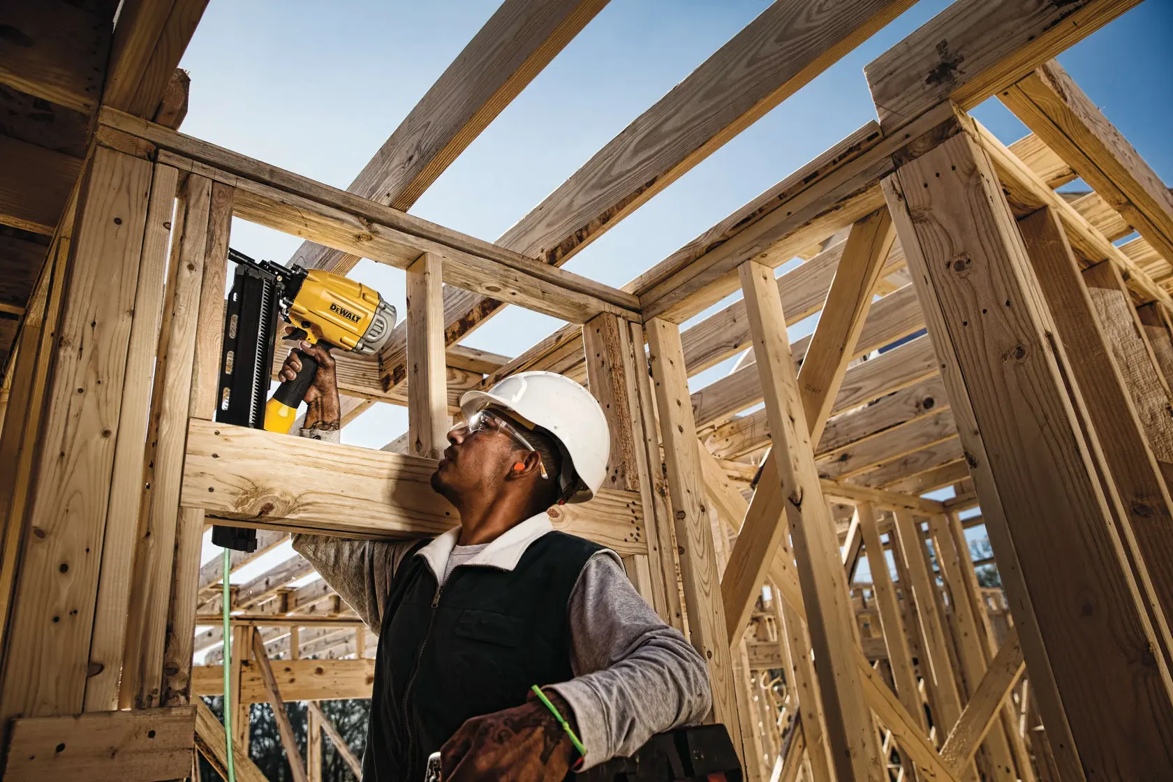 A construction worker wearing a white hard hat uses a yellow DEWALT nail gun to secure wooden beams in a building framework under bright daylight.