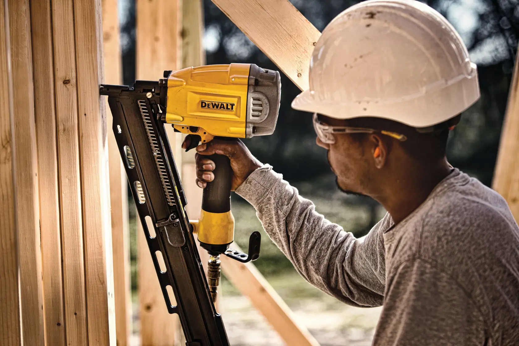 A person wearing a white hard hat uses a DEWALT pneumatic framing nailer to fasten wooden beams at a construction site. The individual’s face is obscured for privacy.