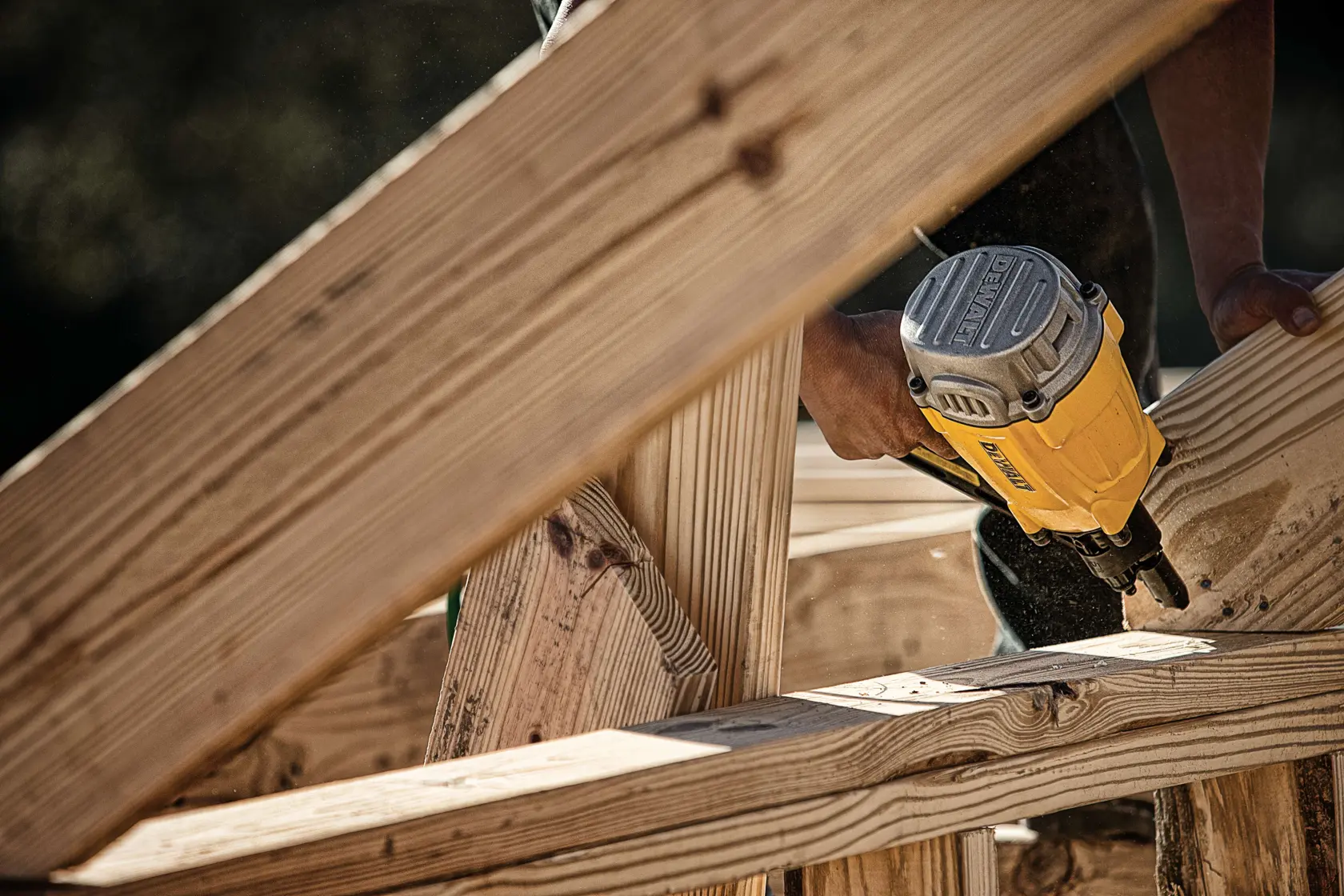 A close-up of a person using a yellow DEWALT nail gun to fasten wooden beams in a construction setting.