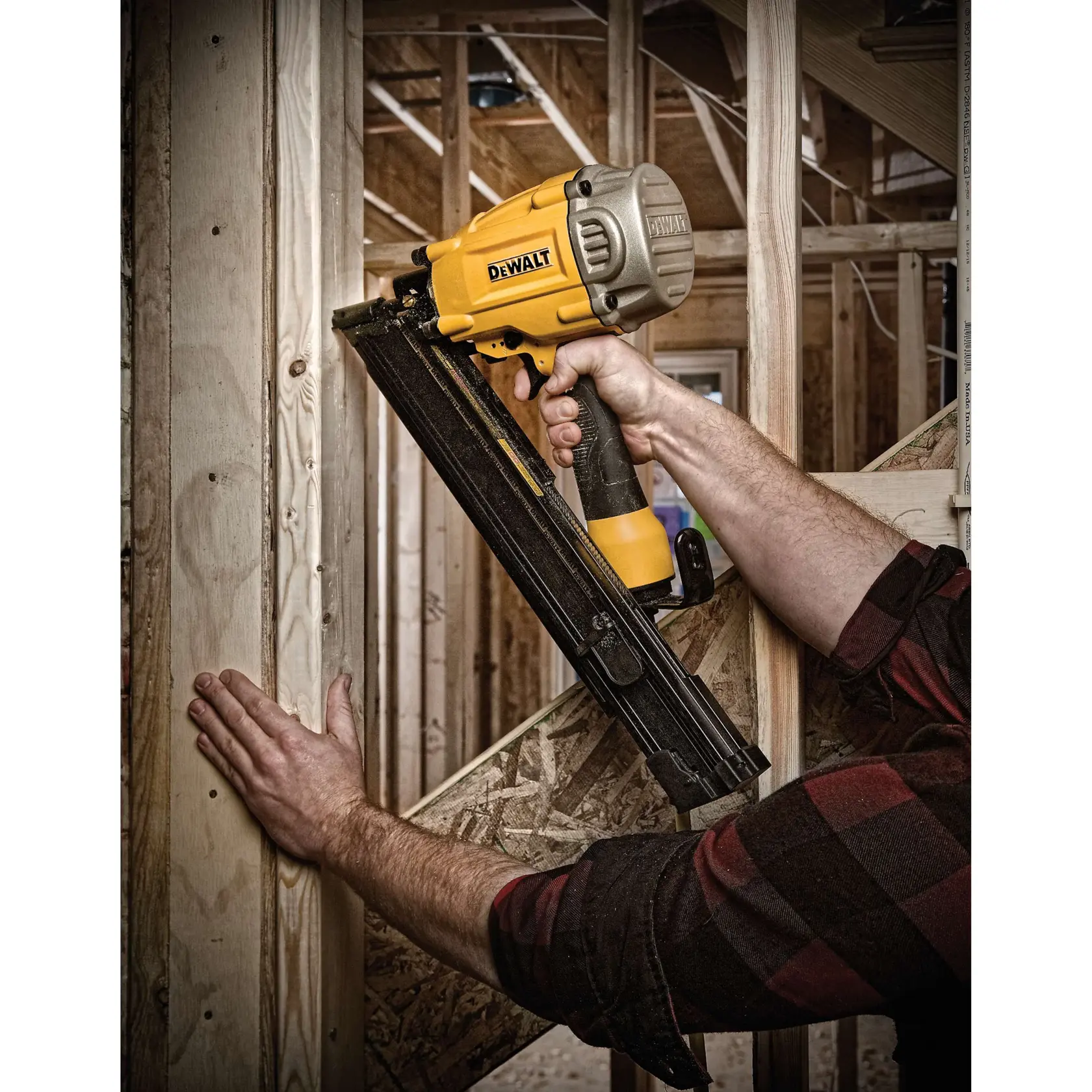 A person is using a DEWALT nail gun to fasten wooden boards in a construction setting. The nail gun is yellow and black and held firmly against the wood framing structure.