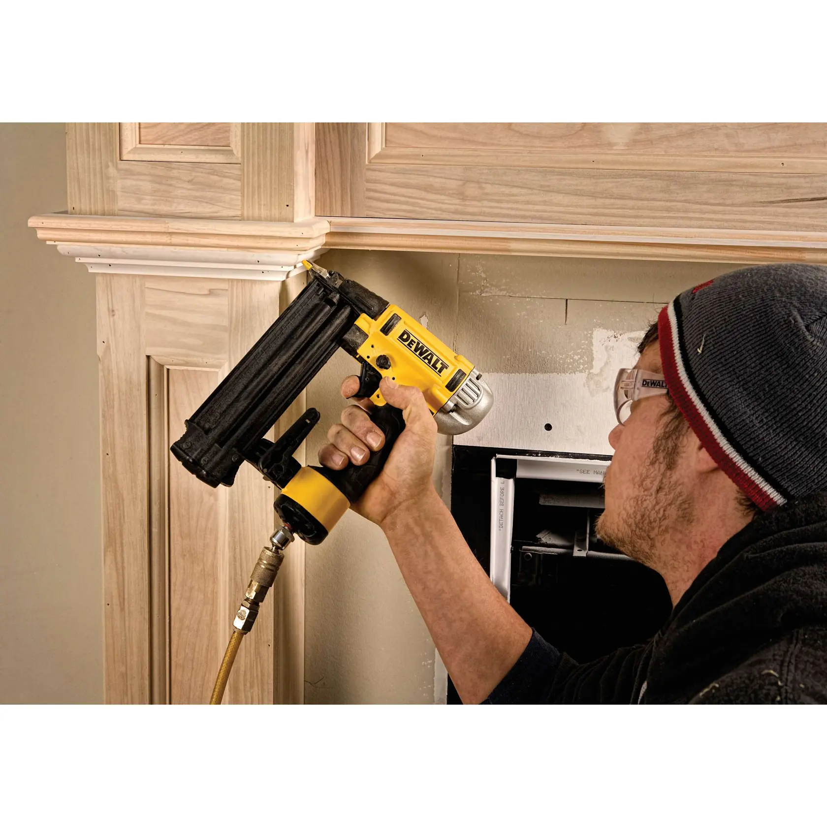 A person is using a DEWALT nail gun to install trim on wooden panels indoors. The nail gun is connected to an air hose. The person is wearing safety glasses and a knit hat.