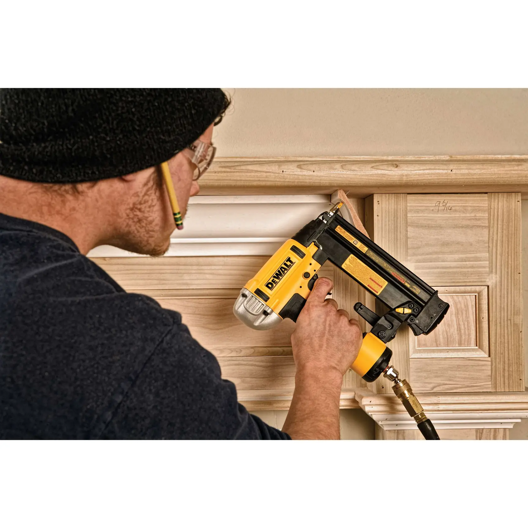 A person is using a DEWALT DWFP12233 nailer to install decorative wood trim on a wall. The person is wearing safety glasses and holding the yellow and black nailer tool against the wood.