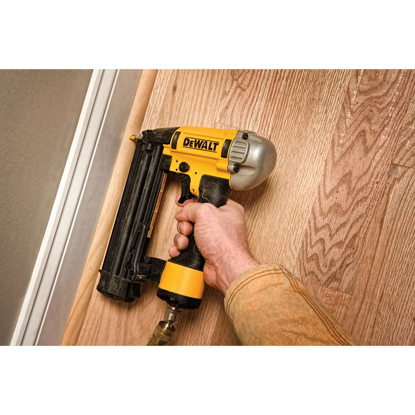 A hand holding a DEWALT pneumatic nailer, model DWFP12233, pressing it against a wooden floor near a baseboard for installation.