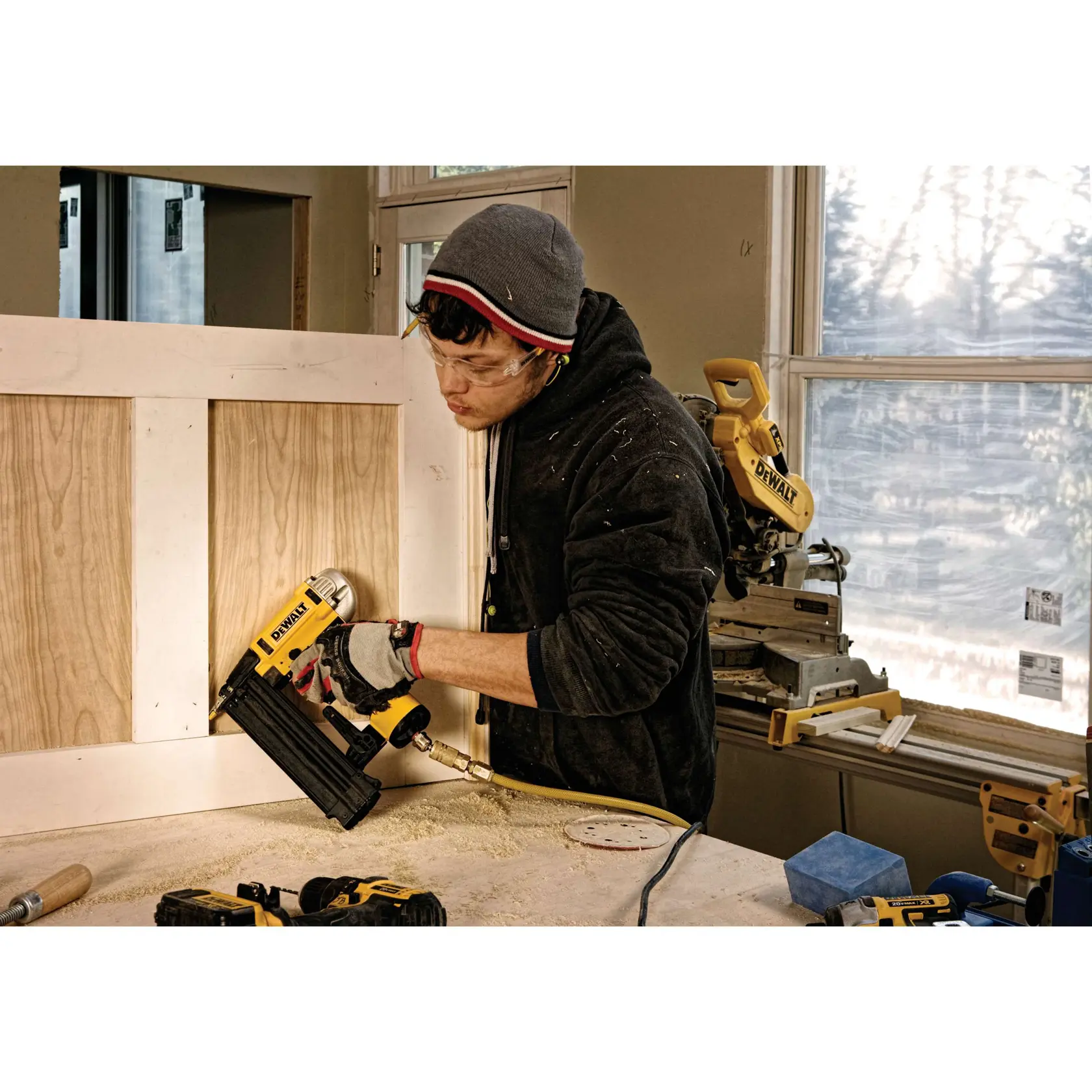 A person wearing safety glasses and gloves is using a DEWALT DWFP12233 pneumatic brad nailer to attach trim to a wooden surface. The image highlights that the tool is ideal for most trim nailing applications and can drive 18-gauge brad nails from 5/8 inch to 2-1/8 inch in length.