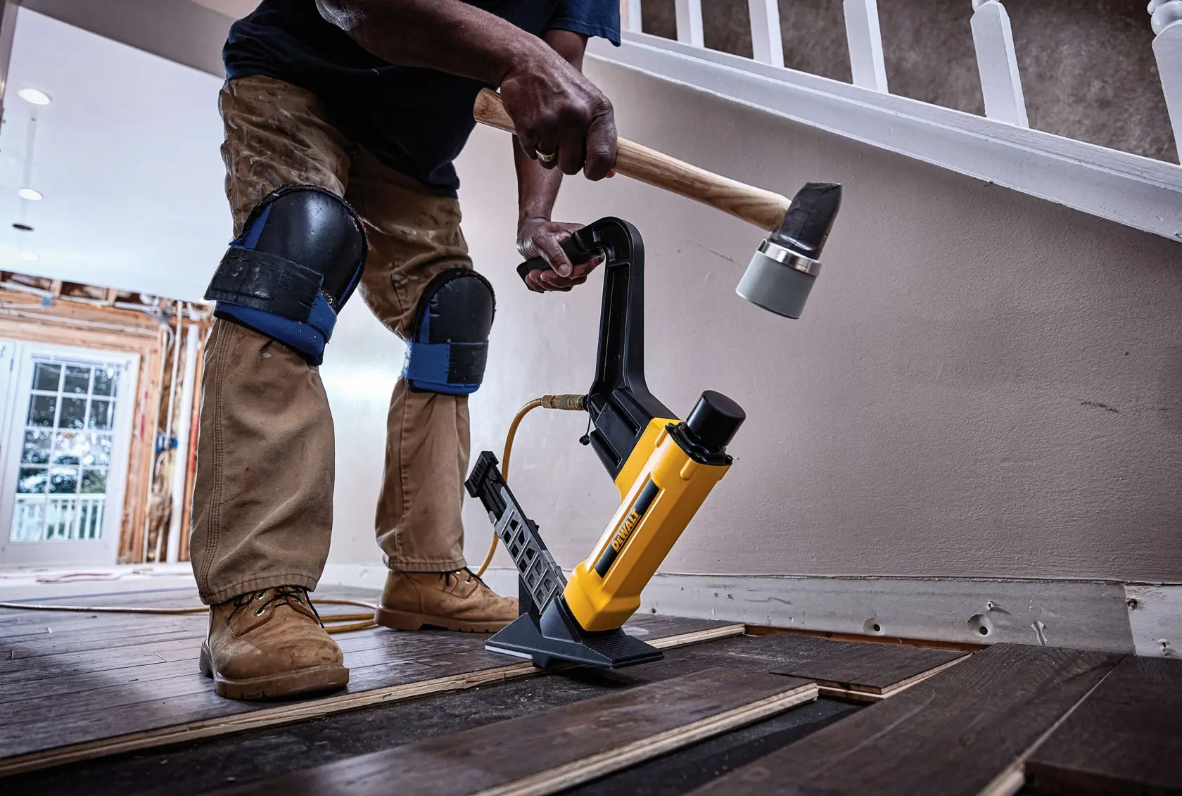A person wearing knee pads is using a DEWALT flooring nailer to install hardwood flooring. The nailer is placed on the wooden planks, and the person is striking it with a mallet. The setting is a partially renovated interior with stairs and a railing in the background.
