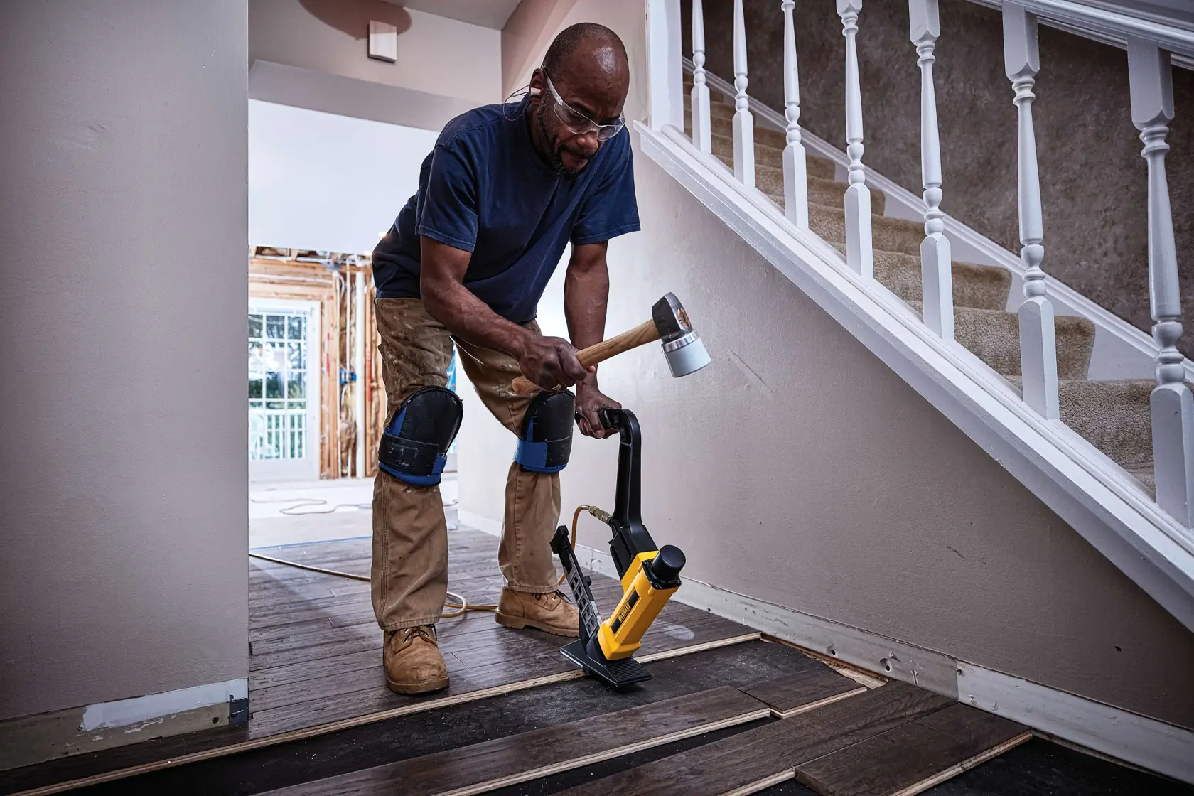 A person wearing knee pads uses a DEWALT flooring nailer and mallet to install wooden floorboards near a staircase inside a house.